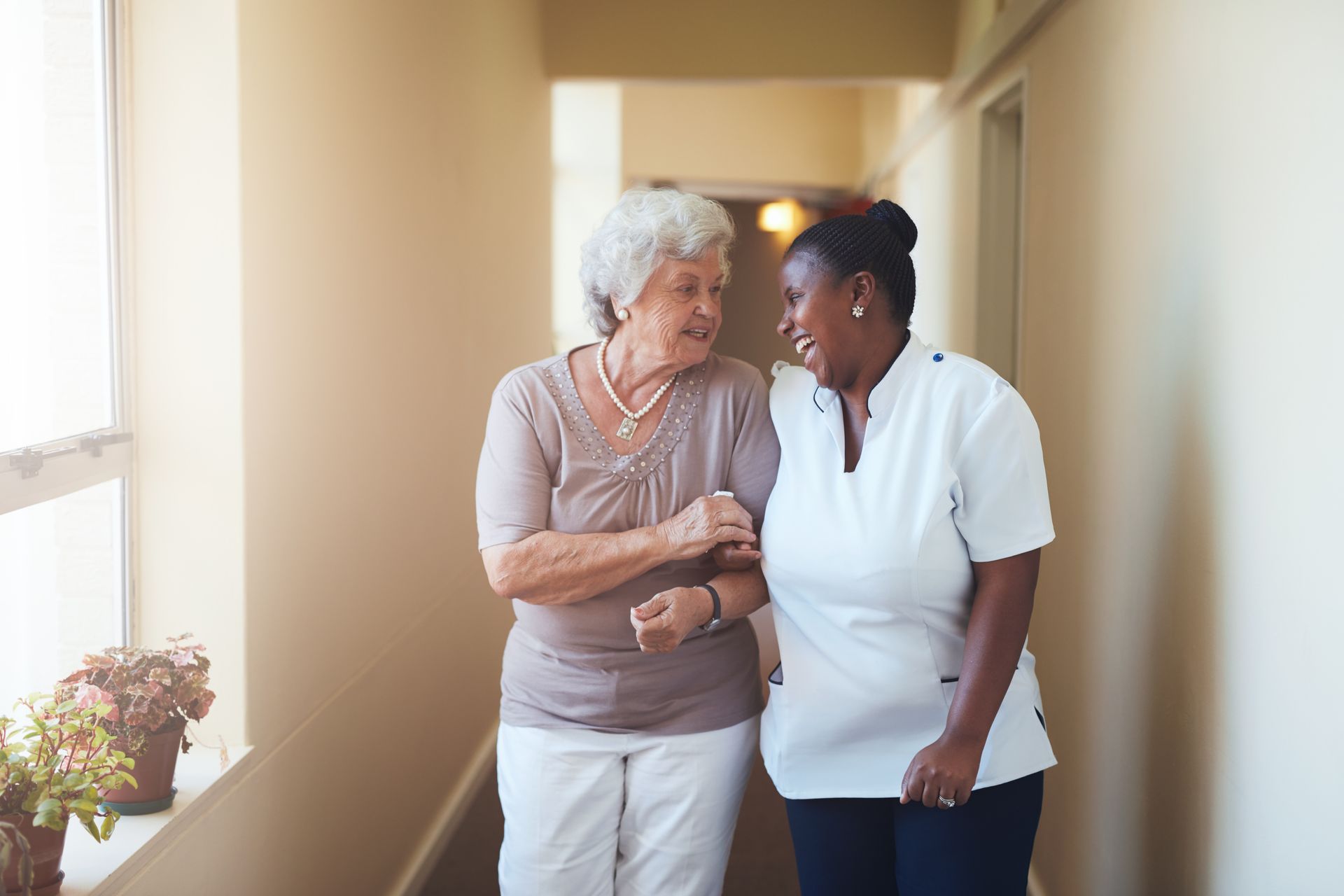 Nurse kneels beside an elderly person in a rocking chair, holding a cane and conversing indoors