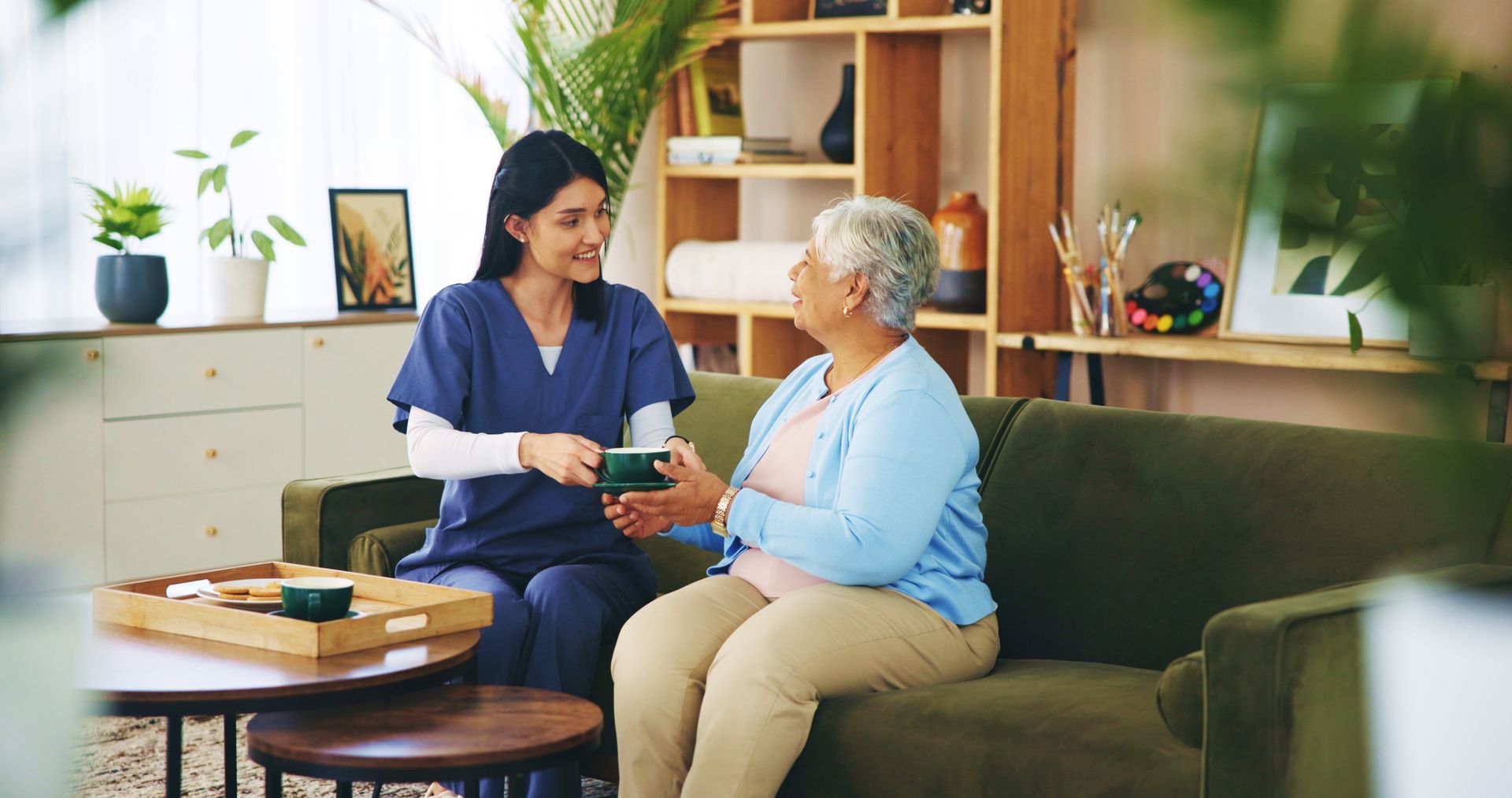 A caregiver in blue scrubs offers a green item to an older person seated on a green couch in a living room