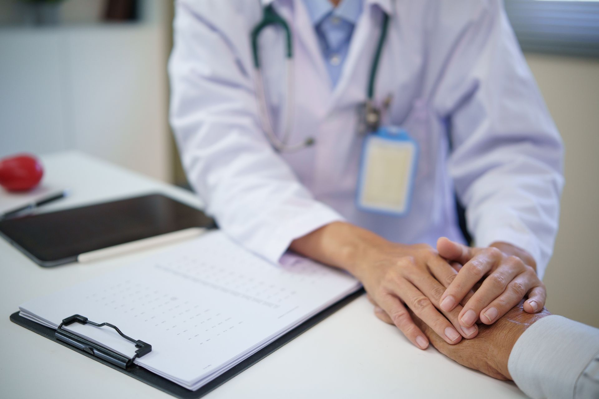 Doctor's hands on a patient's hand; a supportive gesture. White lab coat, stethoscope, clipboard, tablet.