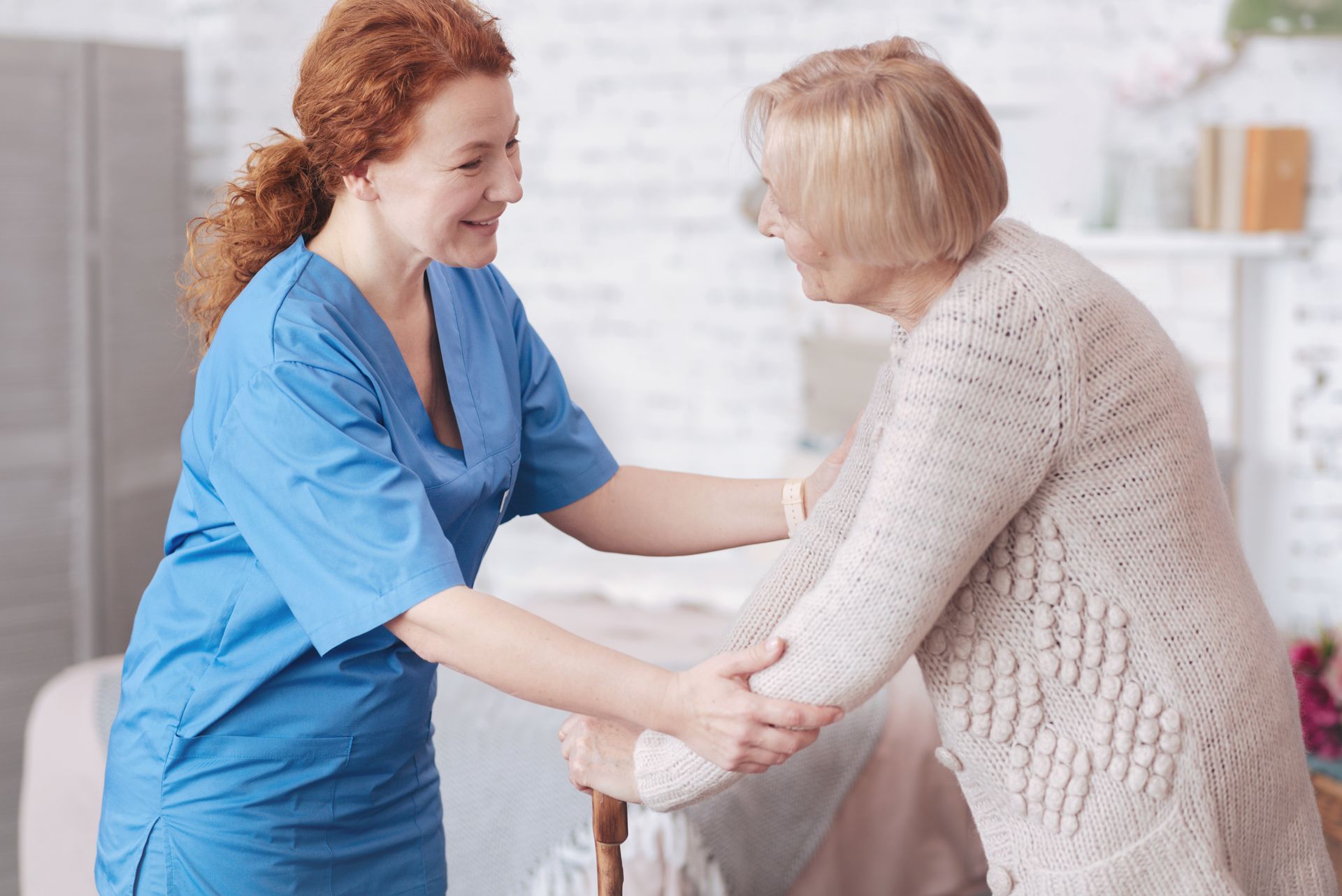 Caregiver in blue scrubs assists an elderly person using a cane; indoors