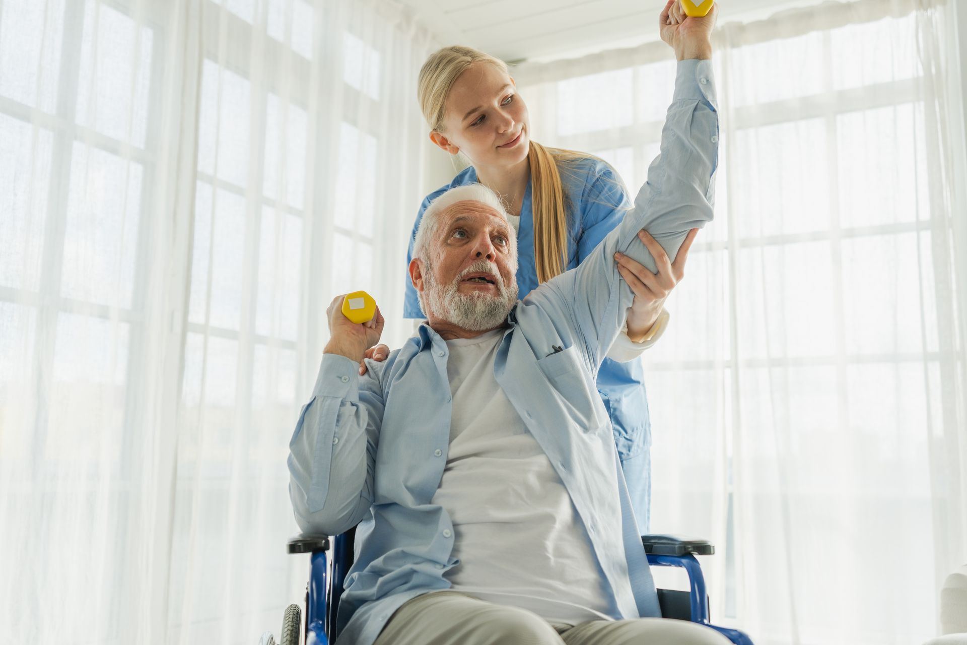 Woman assists seated man with weights, indoors by window Woman assists seated man with weights, indoors by window