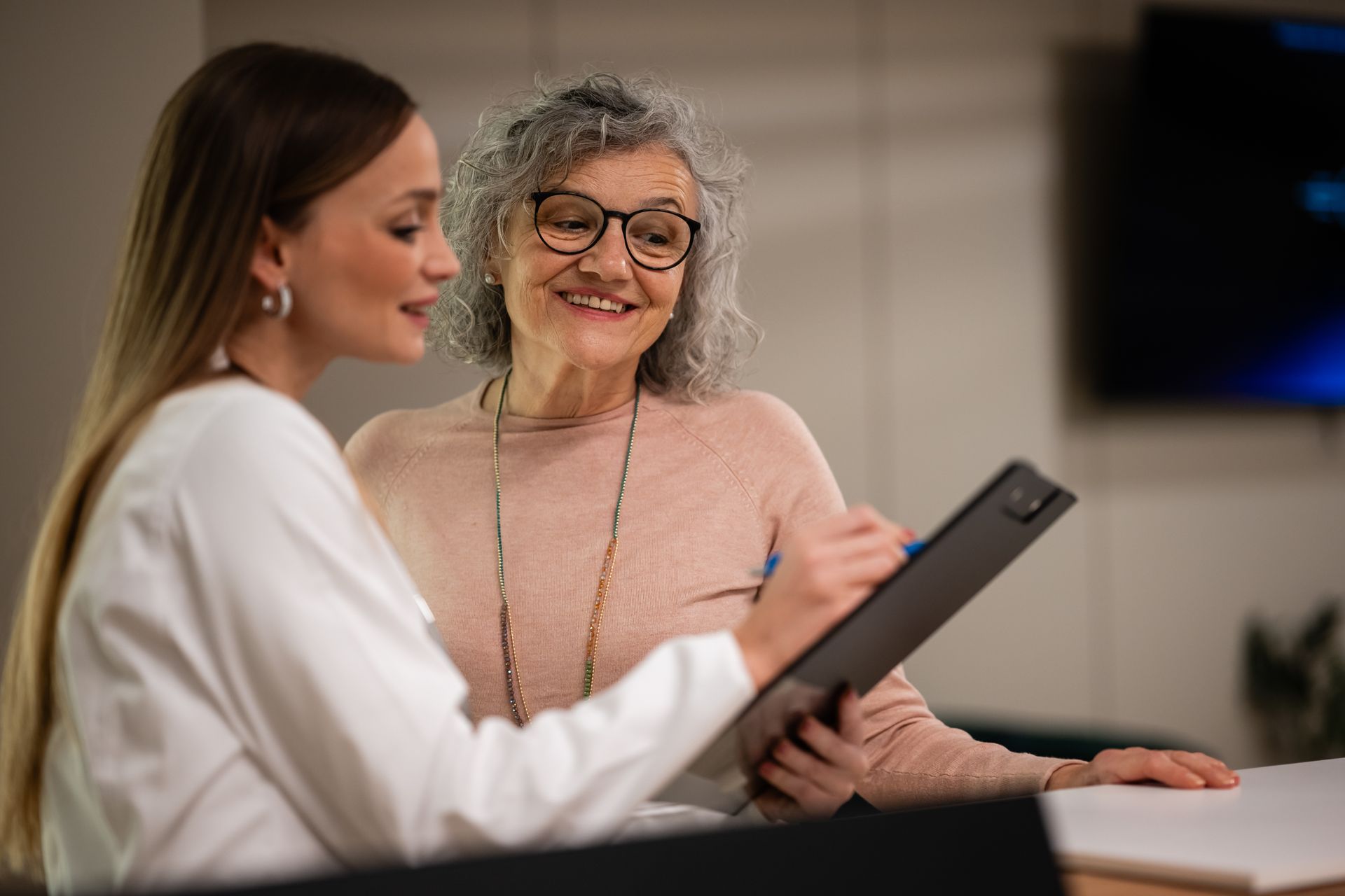 Woman points at document, smiling at older woman wearing glasses. Indoors, neutral tones.