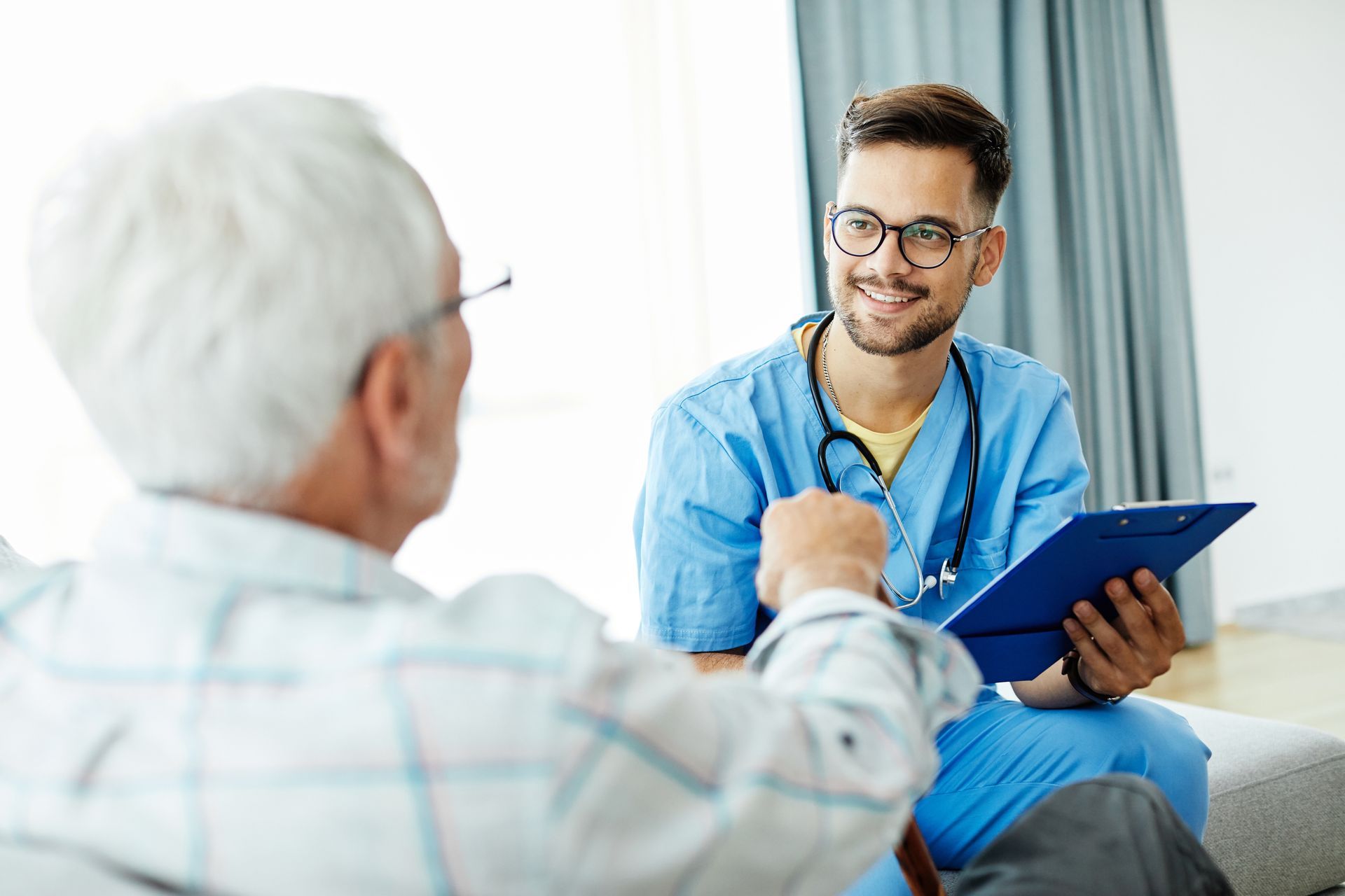 Healthcare worker in blue scrubs smiling while looking at elderly patient, holding clipboard Healthcare worker in blue scrubs smiling while looking at elderly patient, holding clipboard