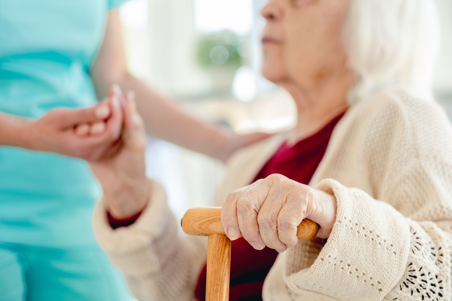 Elderly person holding cane, hand held by person in turquoise scrubs, indoor setting
