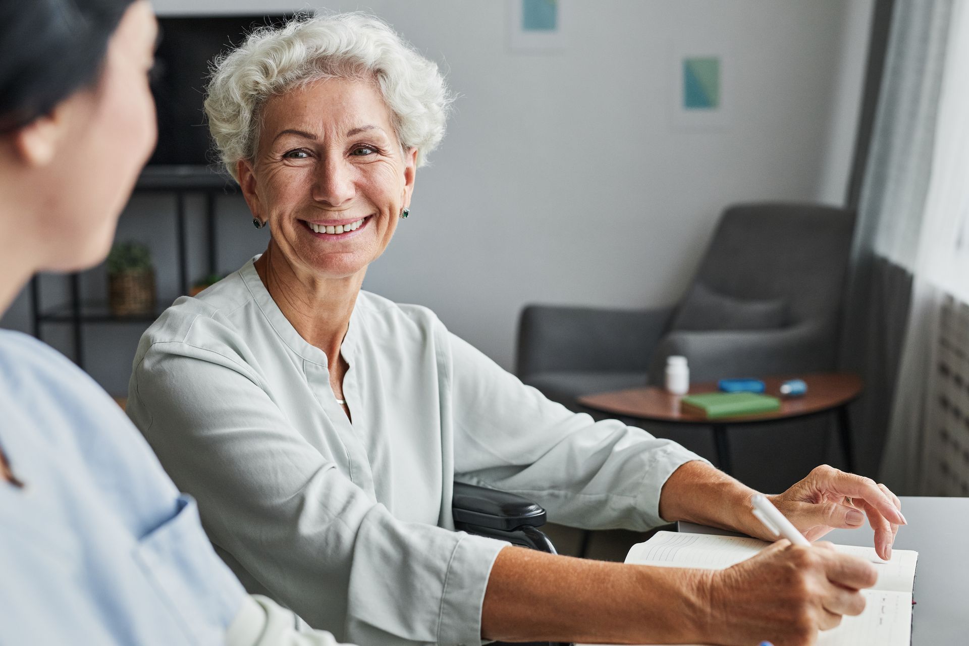 Smiling older adult in a wheelchair writing at a table with a person next to her, indoor setting