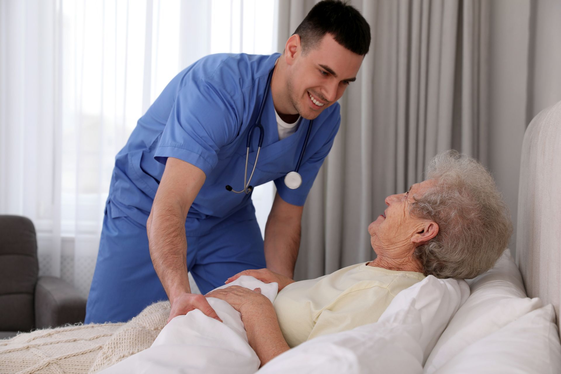 Healthcare worker in blue scrubs assisting an elderly person in bed, smiling and looking down