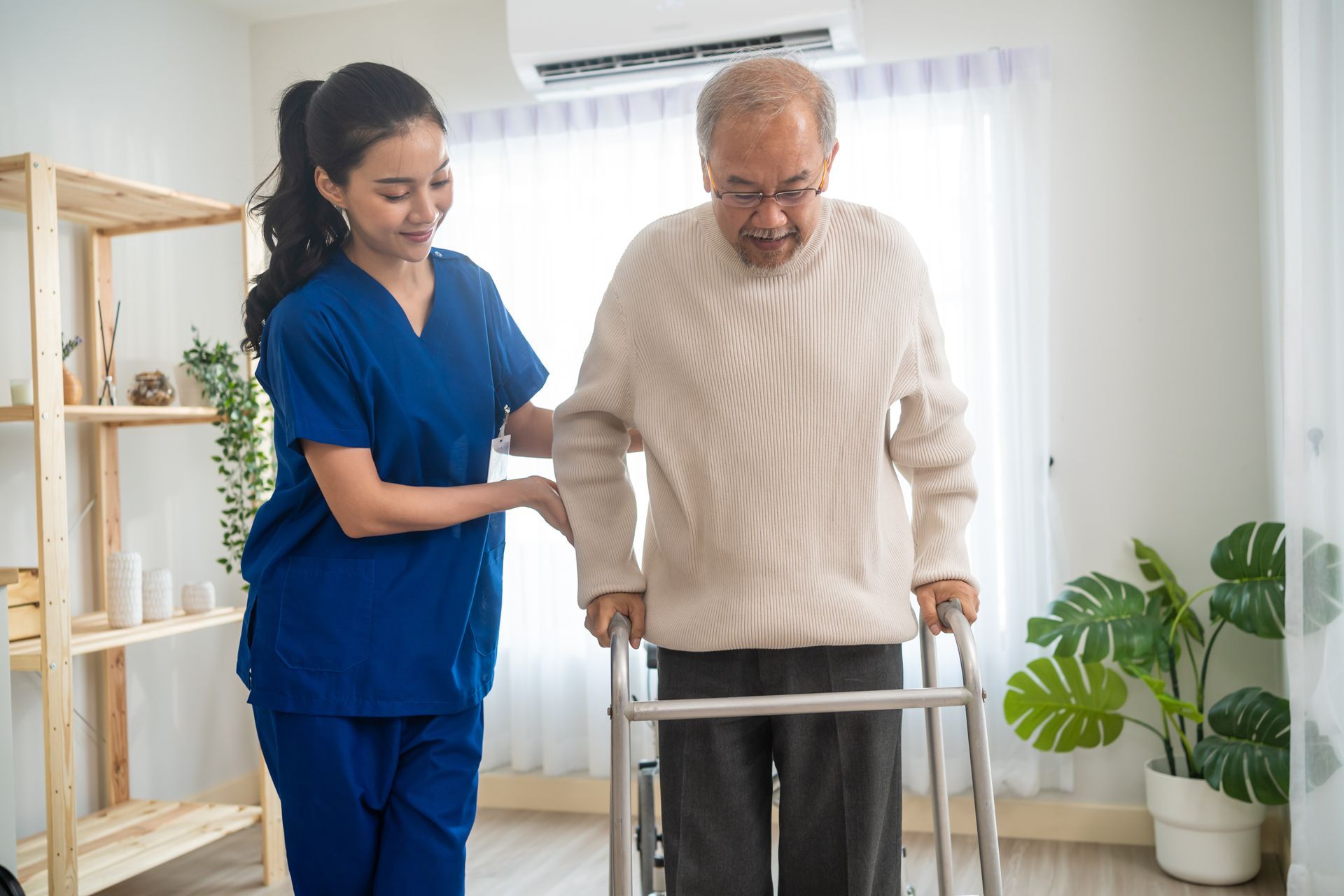 Caregiver assisting a person using a walker in a room; the caregiver wears blue scrubs Caregiver assisting a person using a walker in a room; the caregiver wears blue scrubs