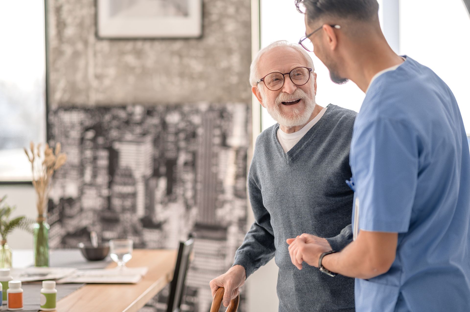 Man with cane smiles, supported by a person in blue scrubs indoors