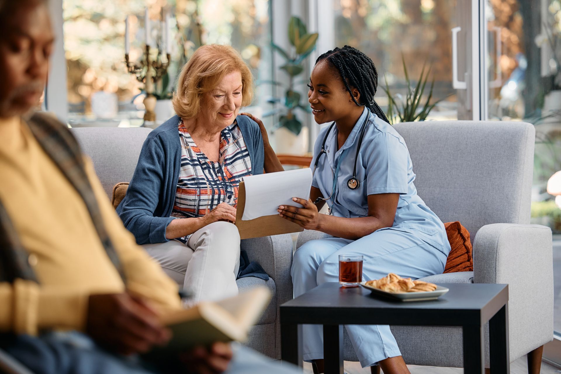 Nurse showing a document to a person seated on a couch; another person reads nearby Nurse showing a document to a person seated on a couch; another person reads nearby