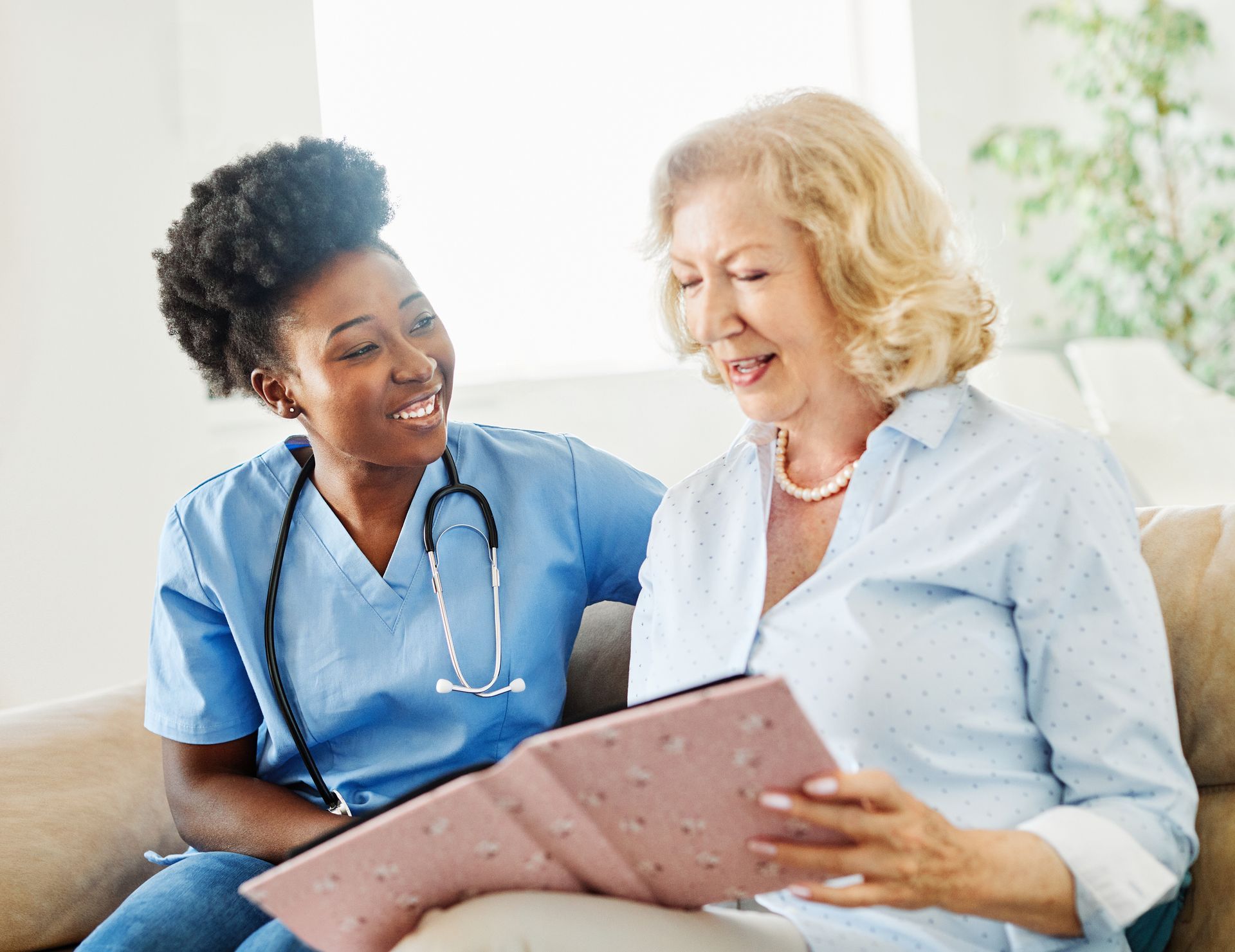 A healthcare worker in blue scrubs smiles at a woman, both looking at a photo album on a sofa A healthcare worker in blue scrubs smiles at a woman, both looking at a photo album on a sofa