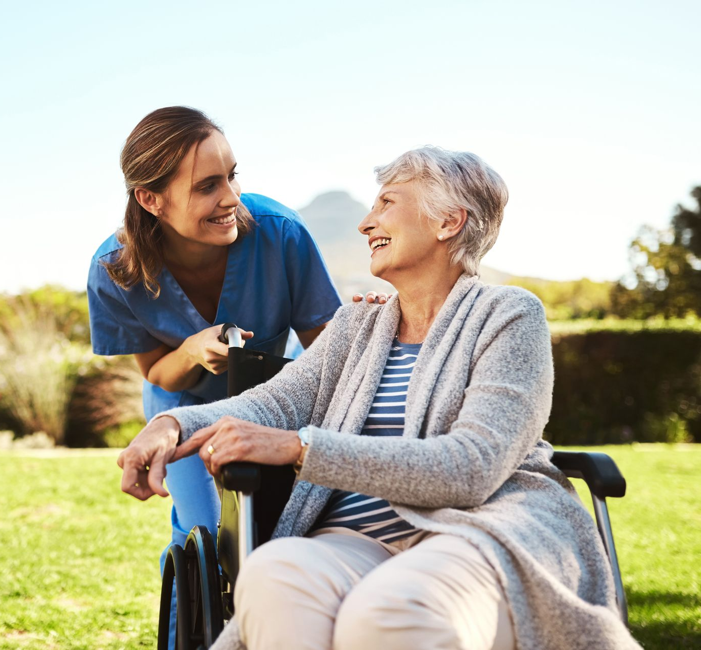 Woman in wheelchair smiles at caregiver in a sunny outdoor setting