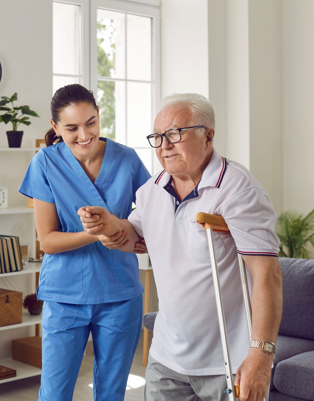 A caregiver assists an elderly person walking with a cane in a bright room