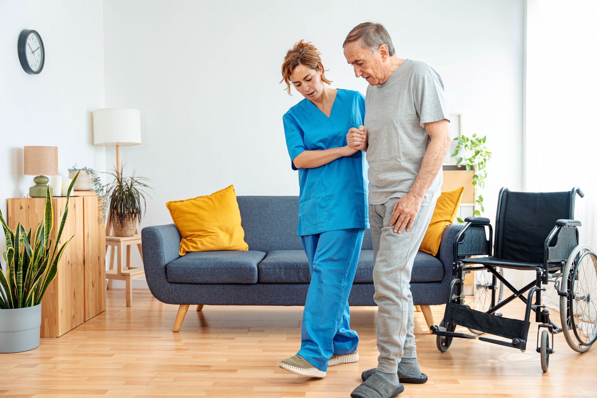 Caregiver assists patient walking; living room setting with wheelchair and sofa Caregiver assists patient walking; living room setting with wheelchair and sofa