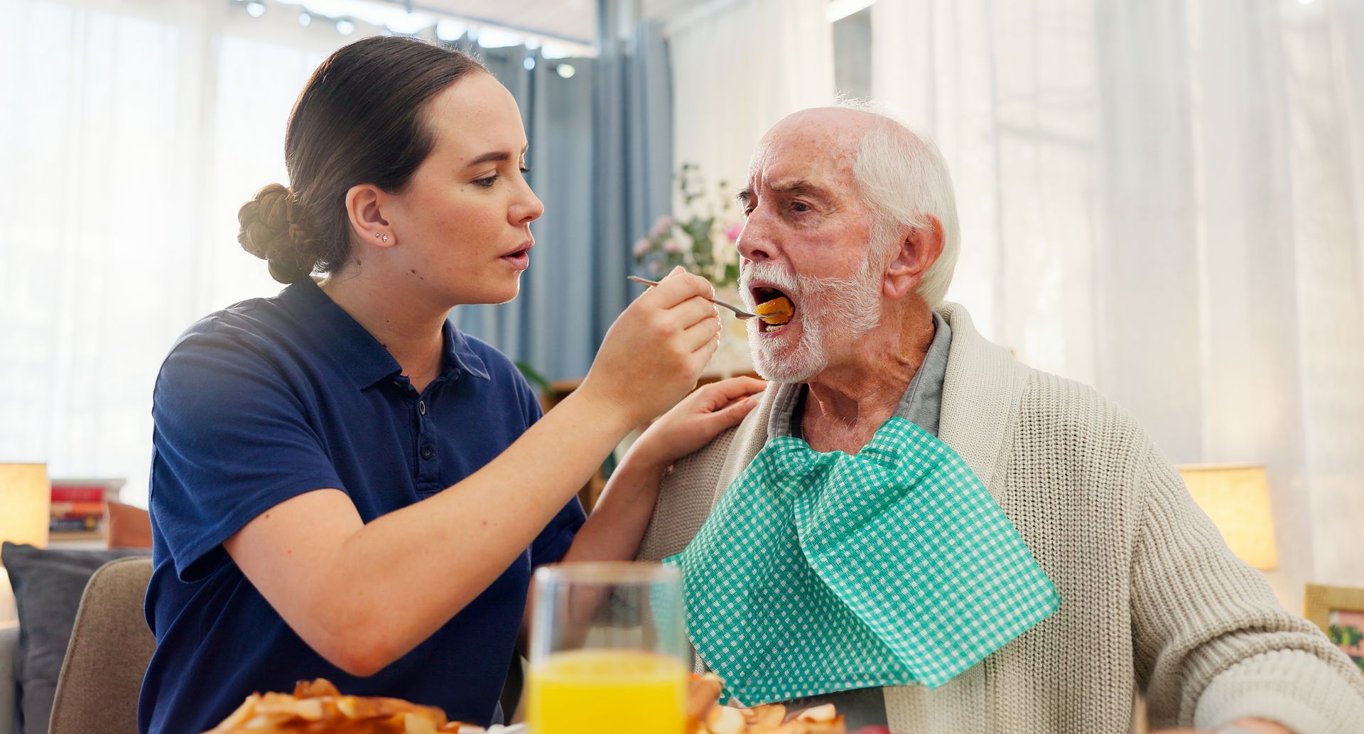 Caregiver feeding an elderly person with a bib in a dining room Caregiver feeding an elderly person with a bib in a dining room