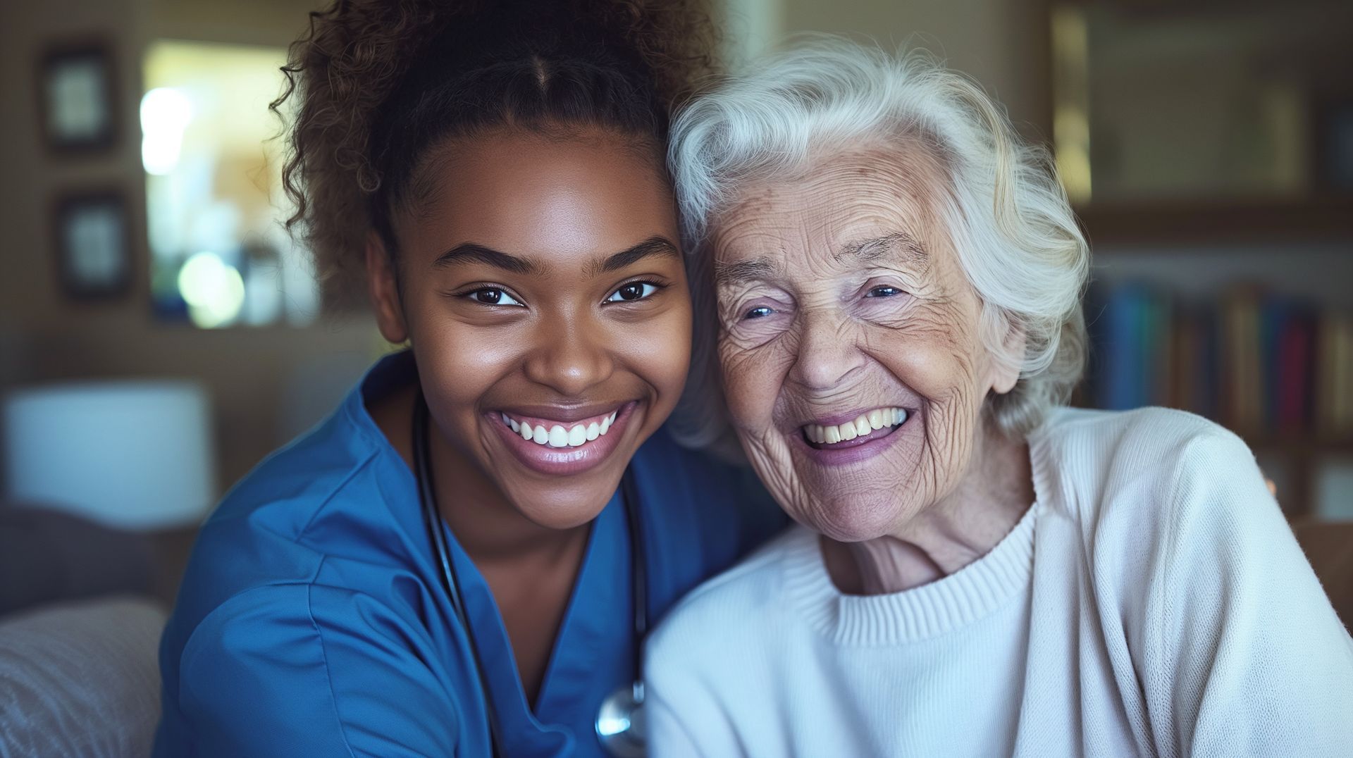 Caregiver in blue scrubs smiles at older person on couch, arms around each other.