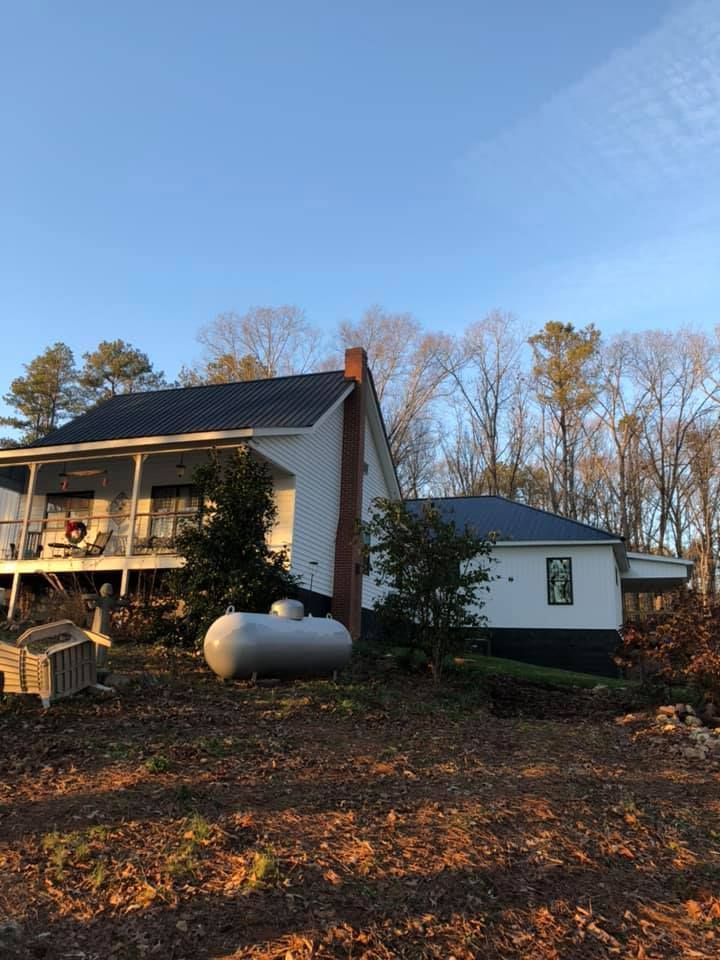 White farmhouse with a dark roof, propane tank, and chimney against a blue sky with bare trees in the background.