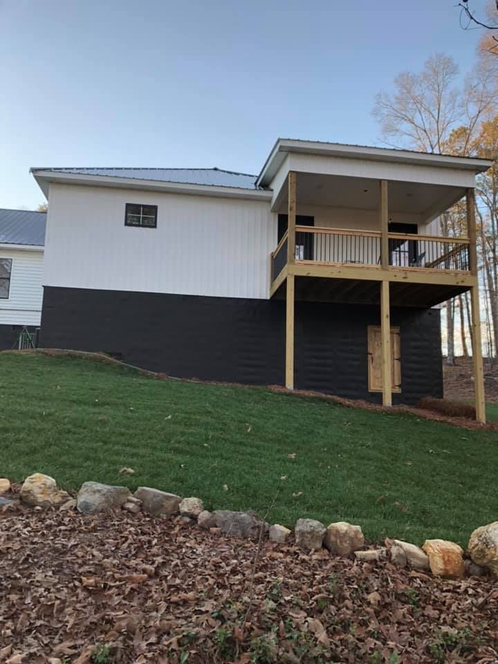 Side view of a two-story house with a dark gray lower level, white upper level, and a covered deck on a grassy hill.