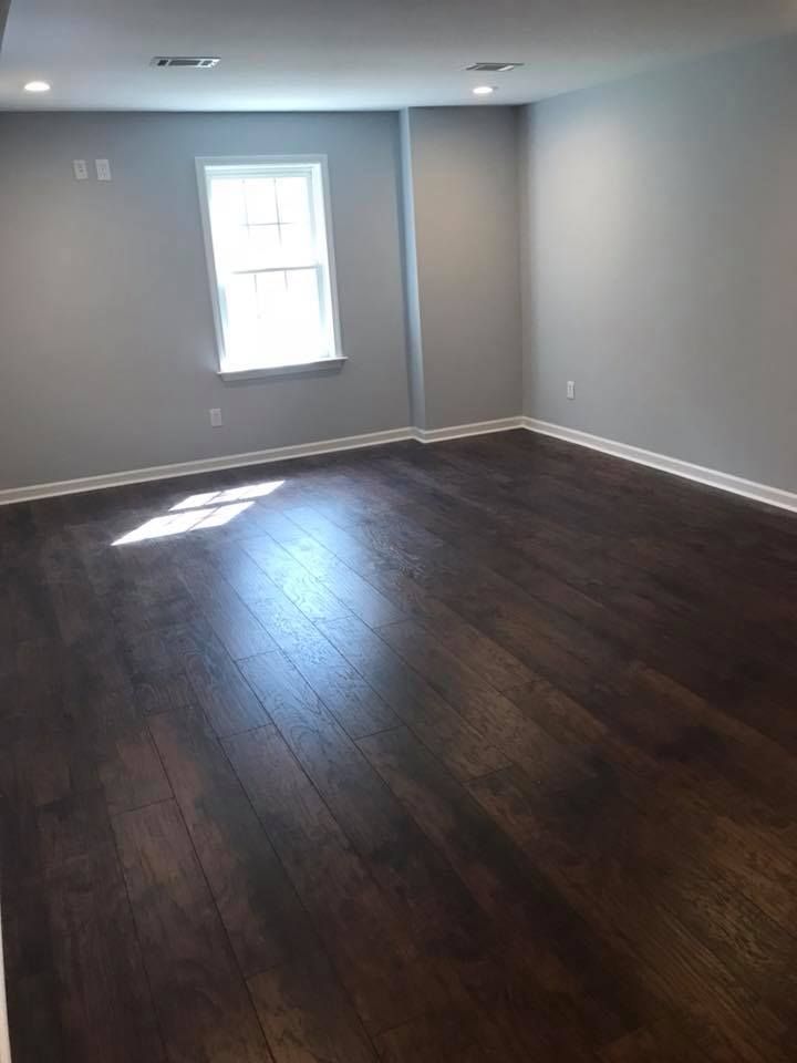 An empty room with dark wood flooring, light gray walls, and a window allowing sunlight.