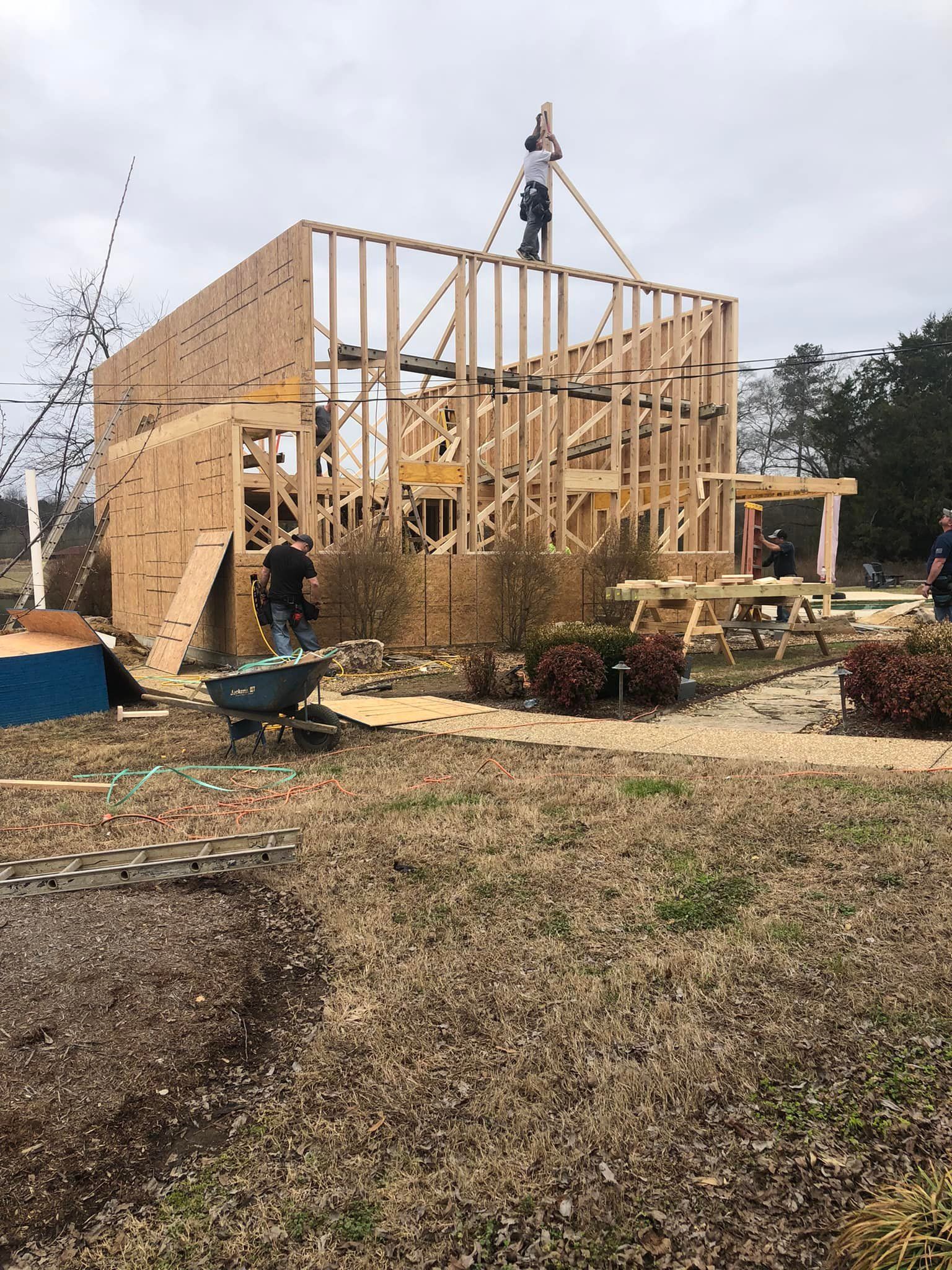 Construction workers framing a two-story wooden structure outdoors, on a cloudy day. One worker atop the frame.