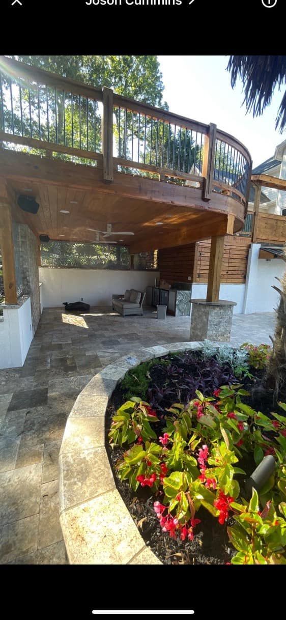 Wooden deck with curved railing overlooking a stone patio and a flower bed with red and green foliage.