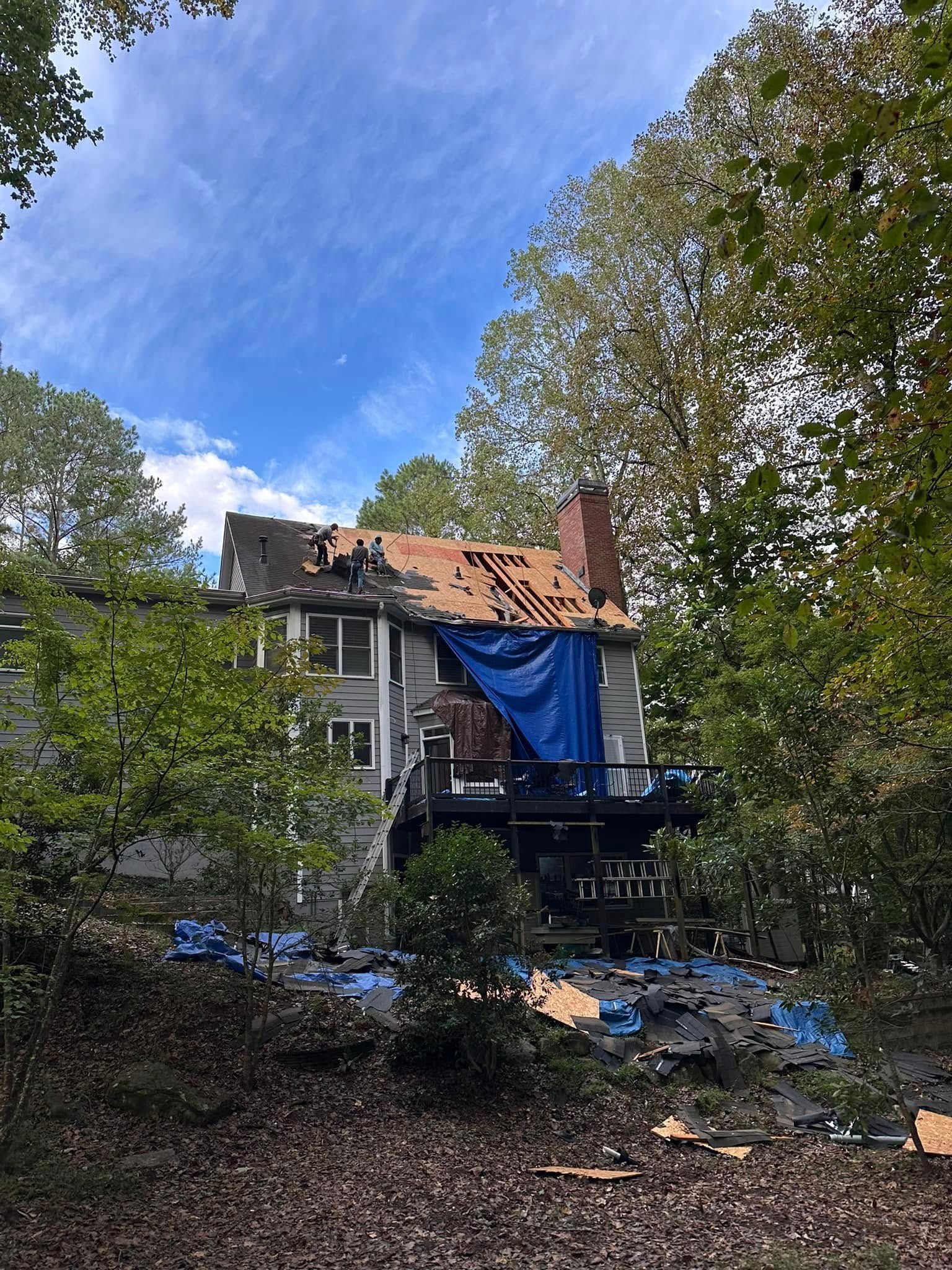 House with partially removed roof covered by a blue tarp, surrounded by trees under a blue sky.