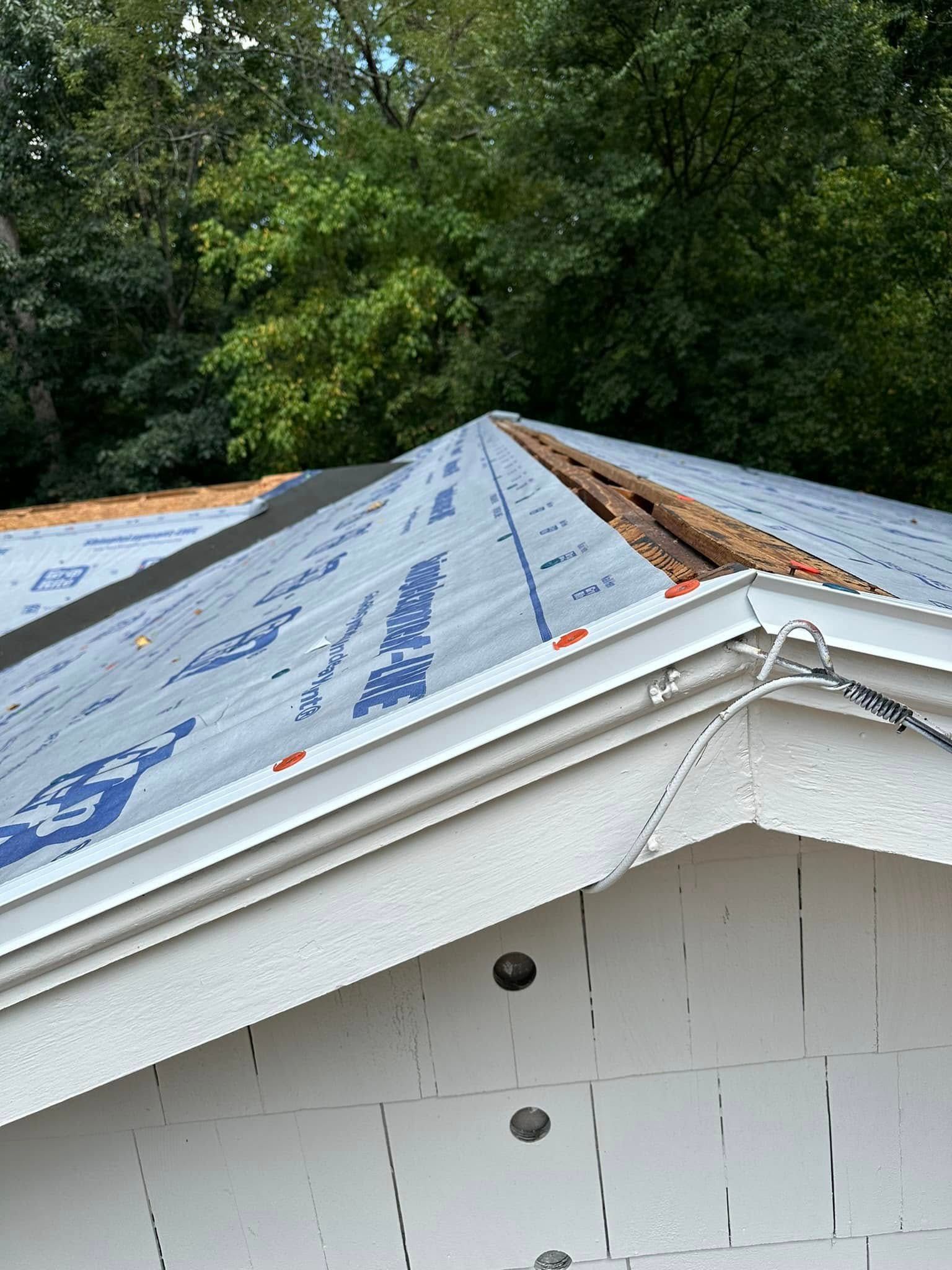 A roof partially covered with blue underlayment, white trim, and exposed wood at the ridge,