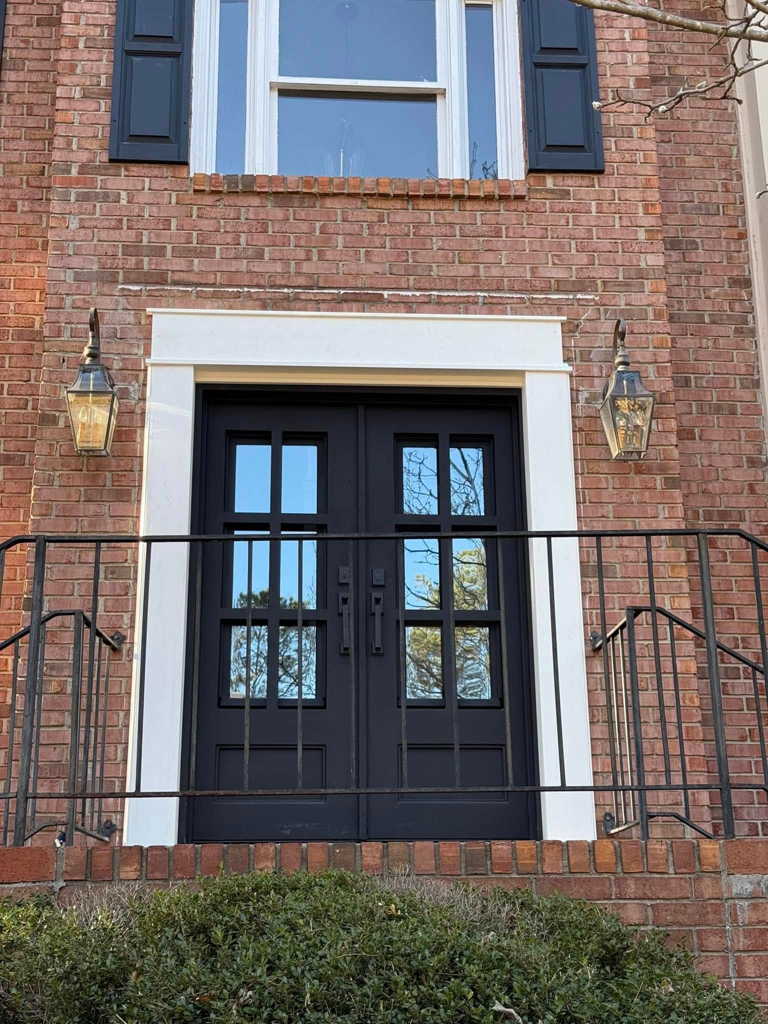 Black double doors with glass panes on a red brick building, flanked by sconces and railing. White trim around the door.