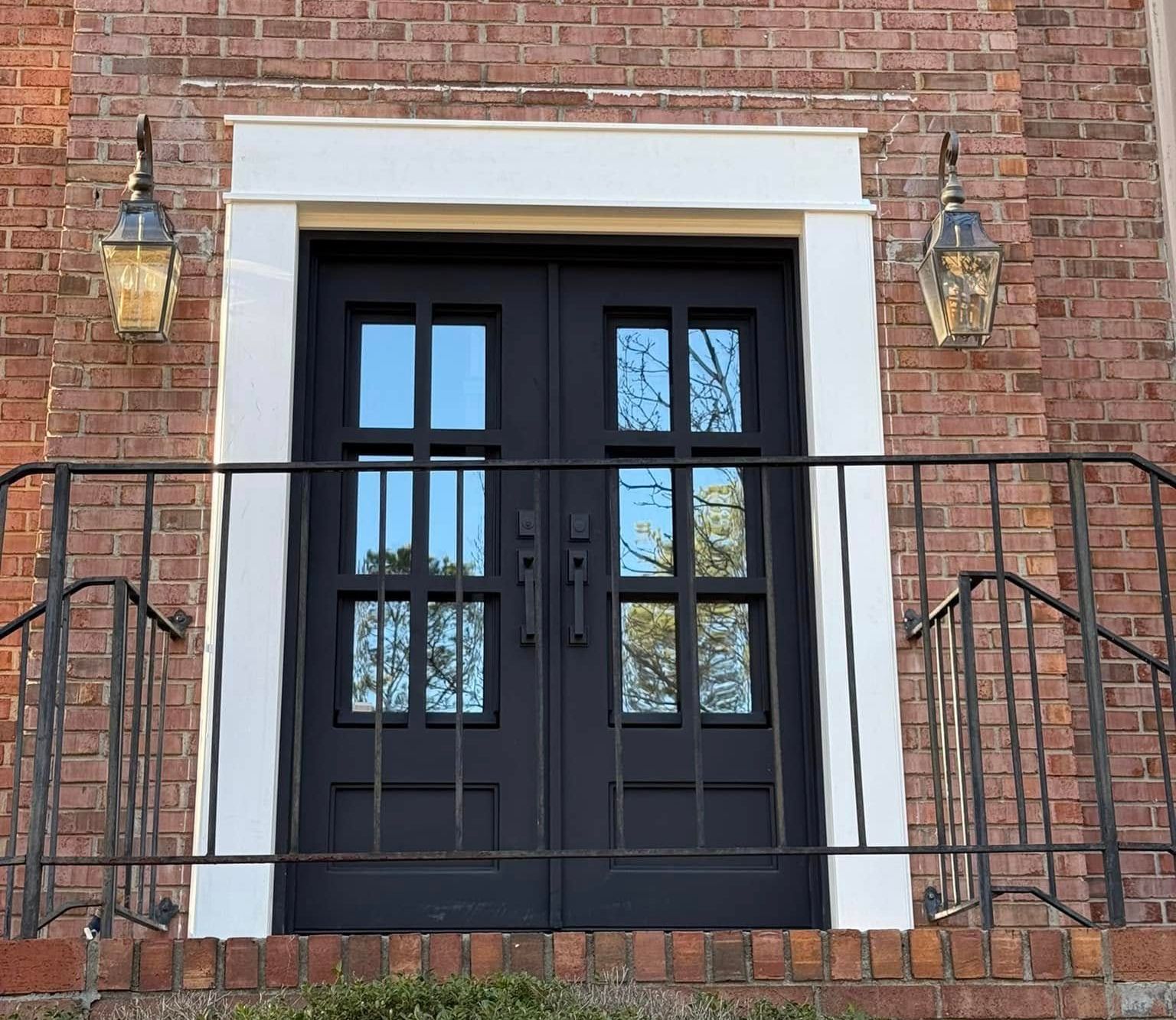 Black double doors with glass panes on a red brick building, flanked by sconces and railing. White trim around the door.
