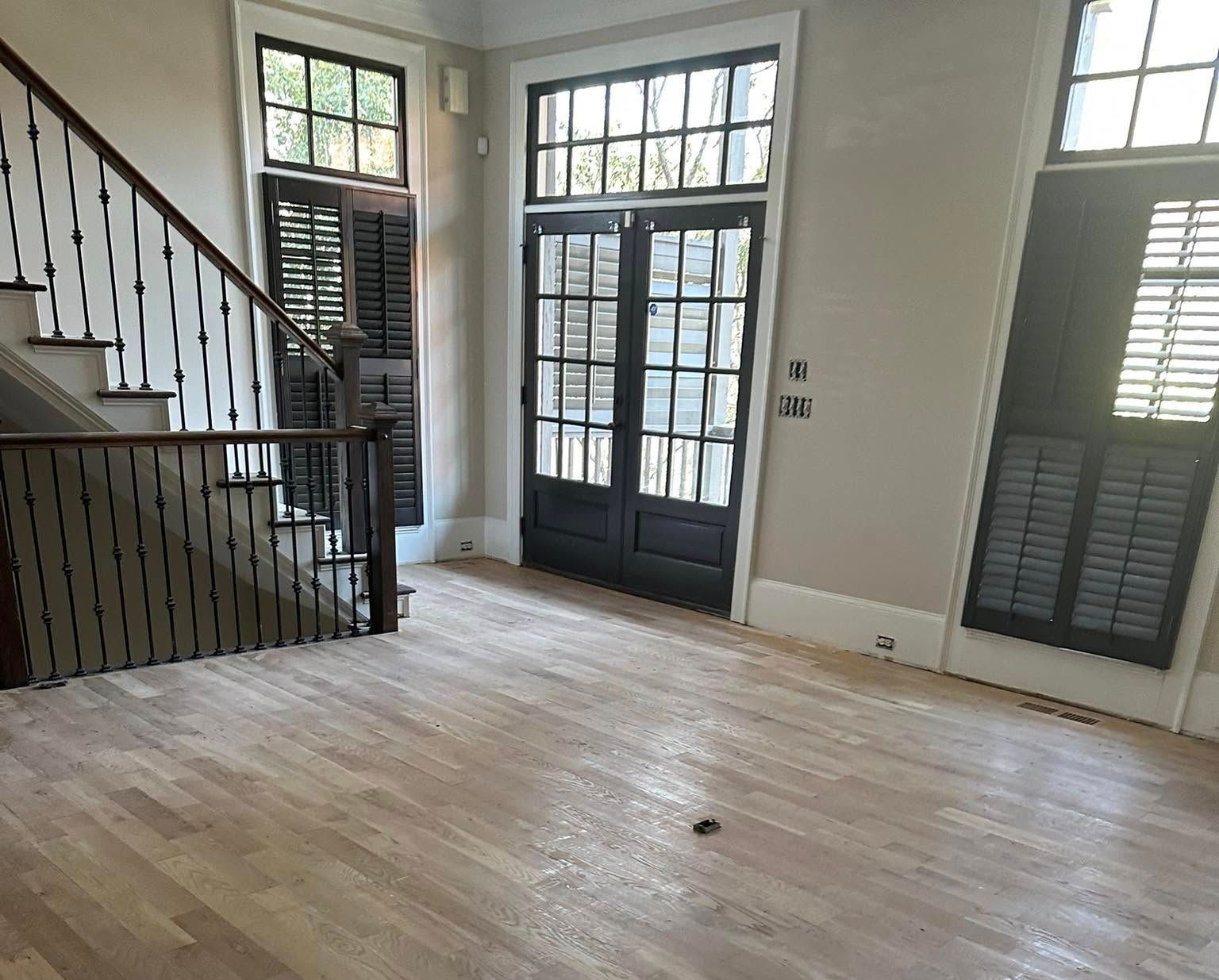 Wooden floor, staircase, and doors with shutters in a mostly empty room with light gray walls.