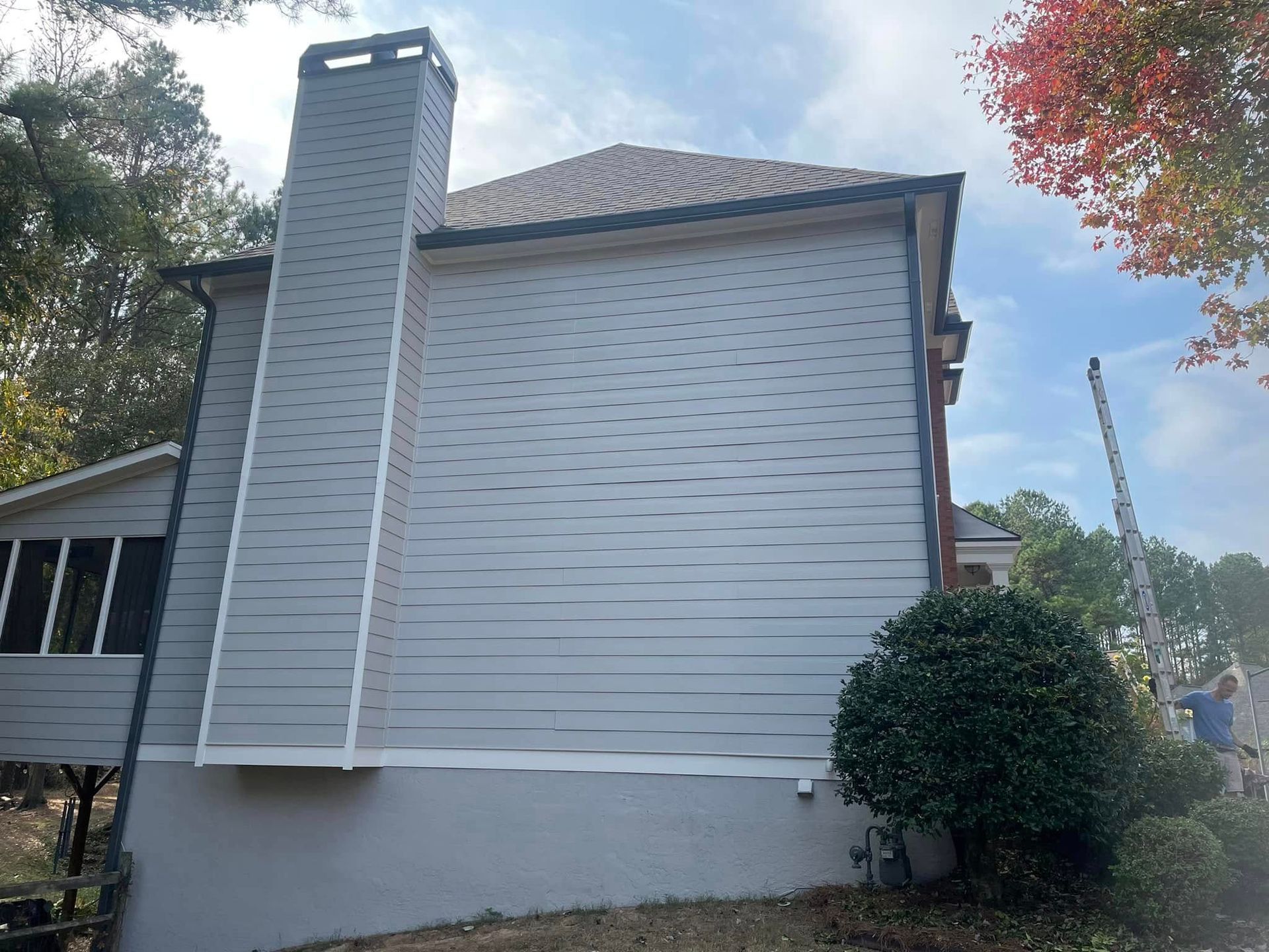 Side view of a house with gray siding and a tall chimney. A porch and trees are visible in the background.