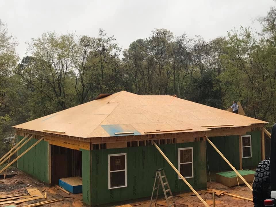 House under construction with green siding and plywood roof, supported by bracing; set in a wooded area.