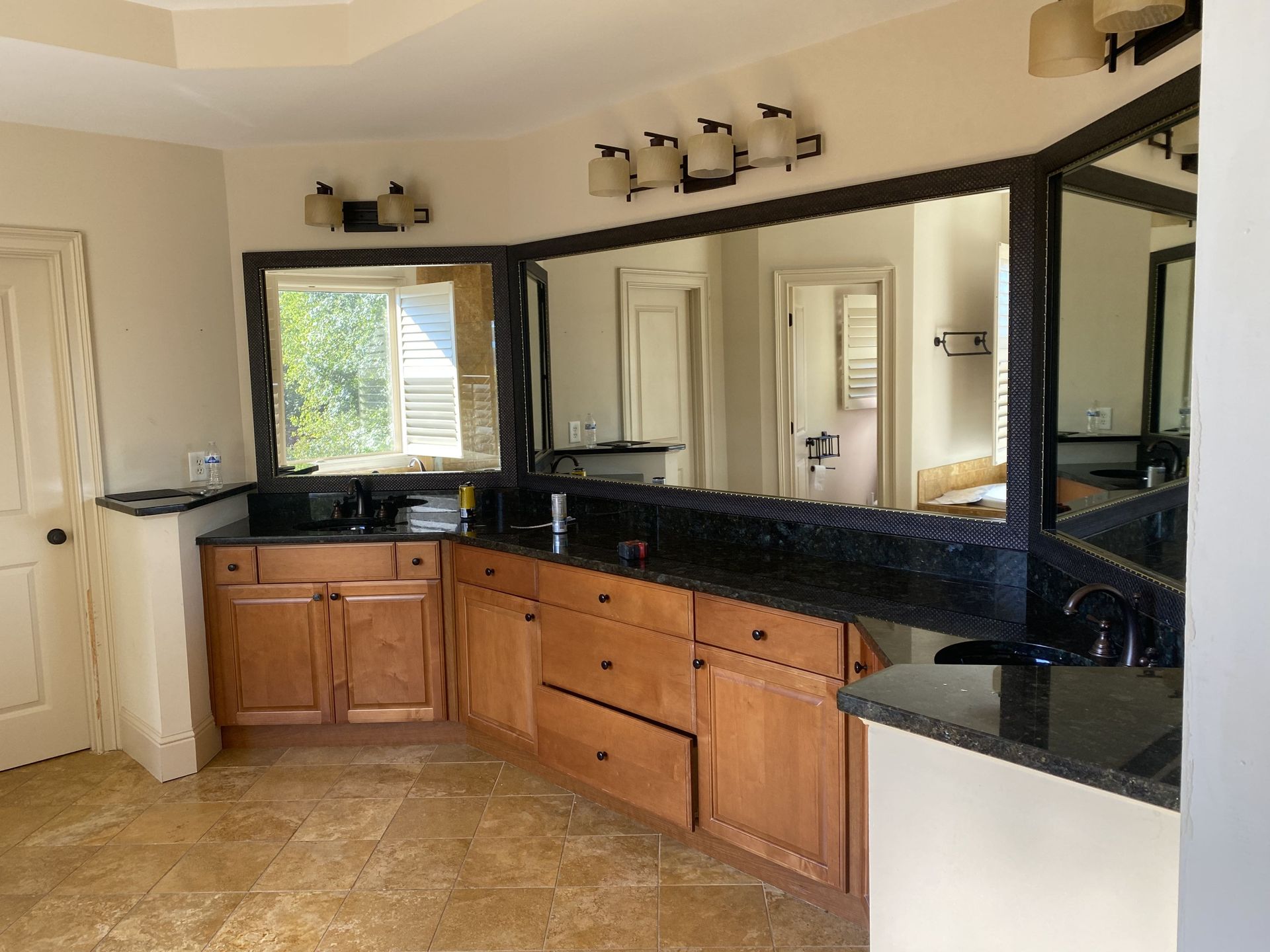 Bathroom with light wood cabinets, black countertops, large mirrors, and beige tiled floor.