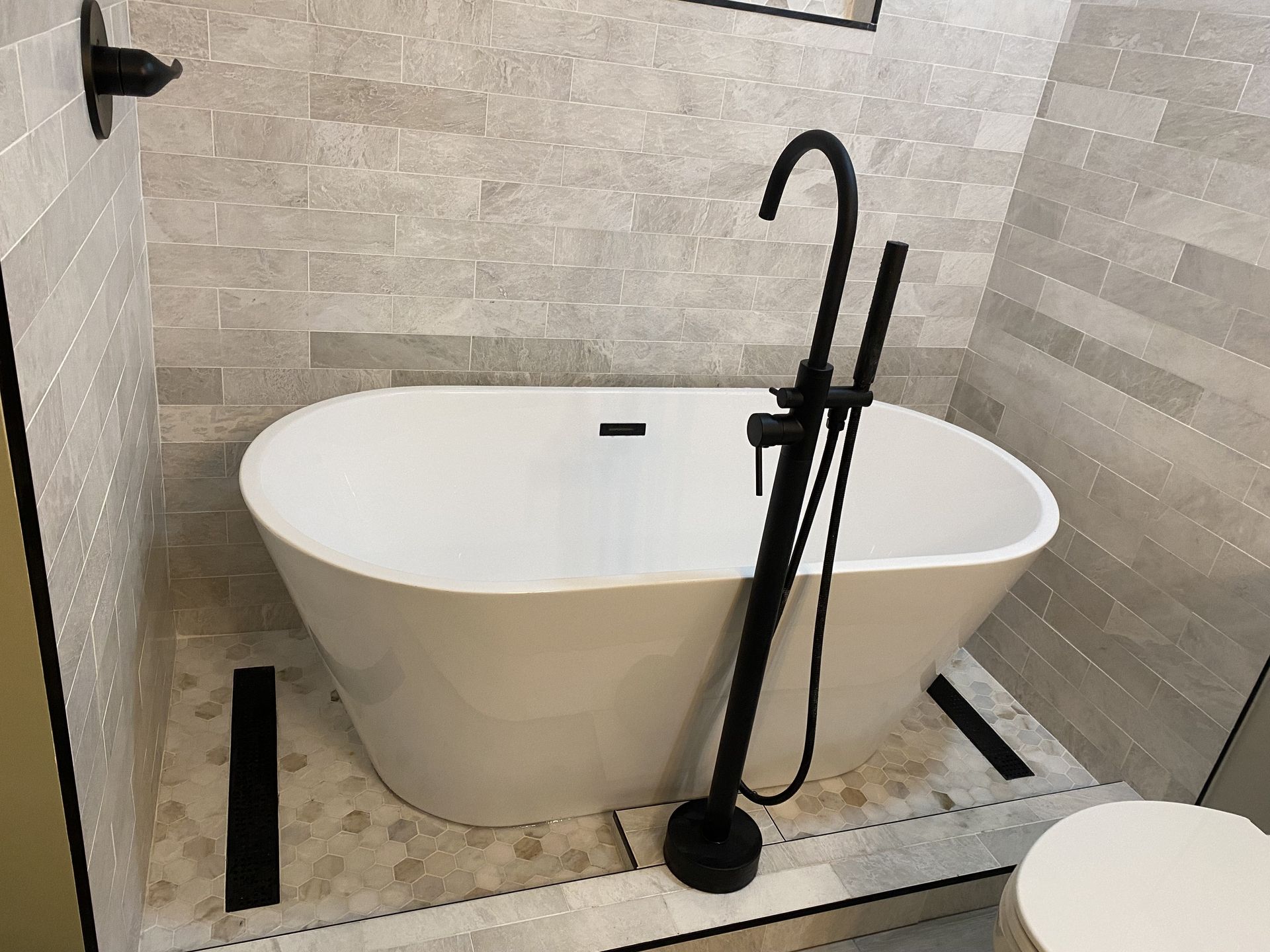 Freestanding white bathtub with black faucet in a gray-tiled bathroom.