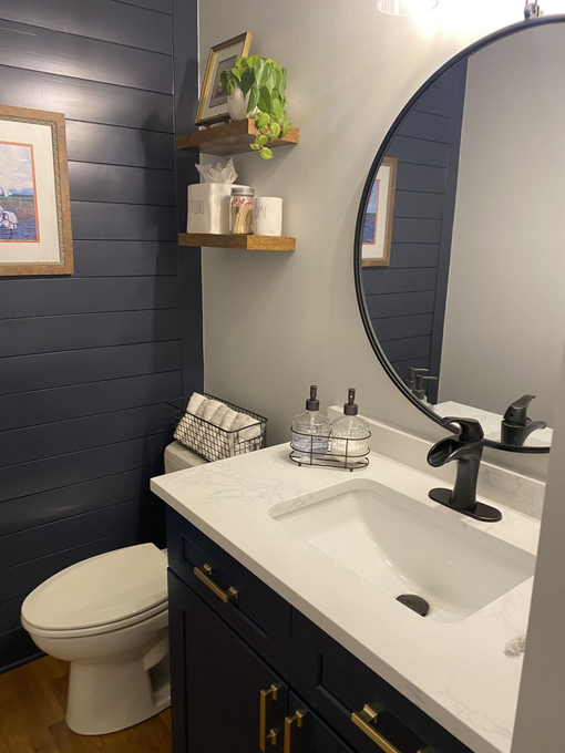Navy blue and gray bathroom with white vanity, round mirror, and wood shelving.