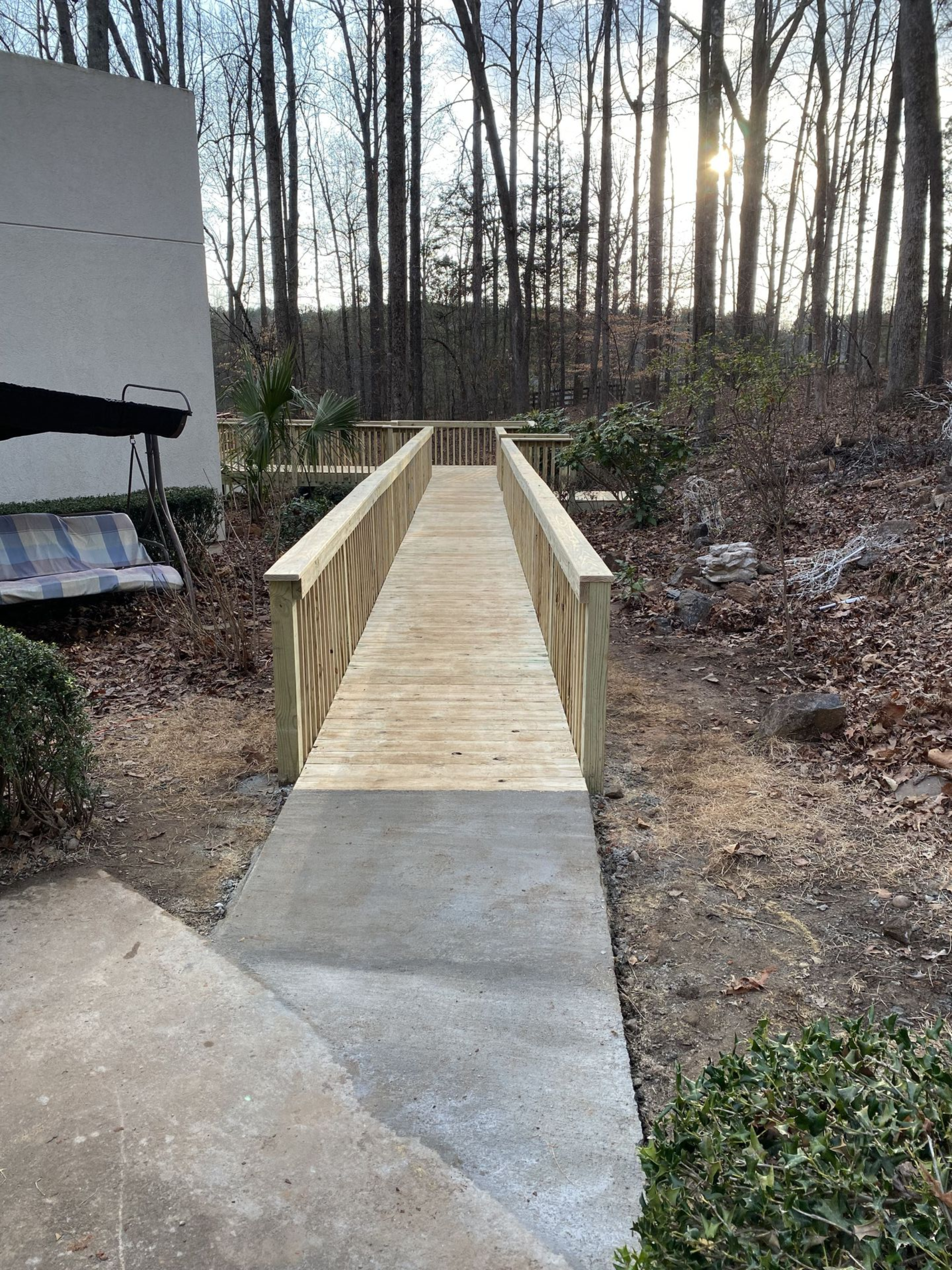 Wooden ramp with handrails extends from a concrete path into a wooded area.