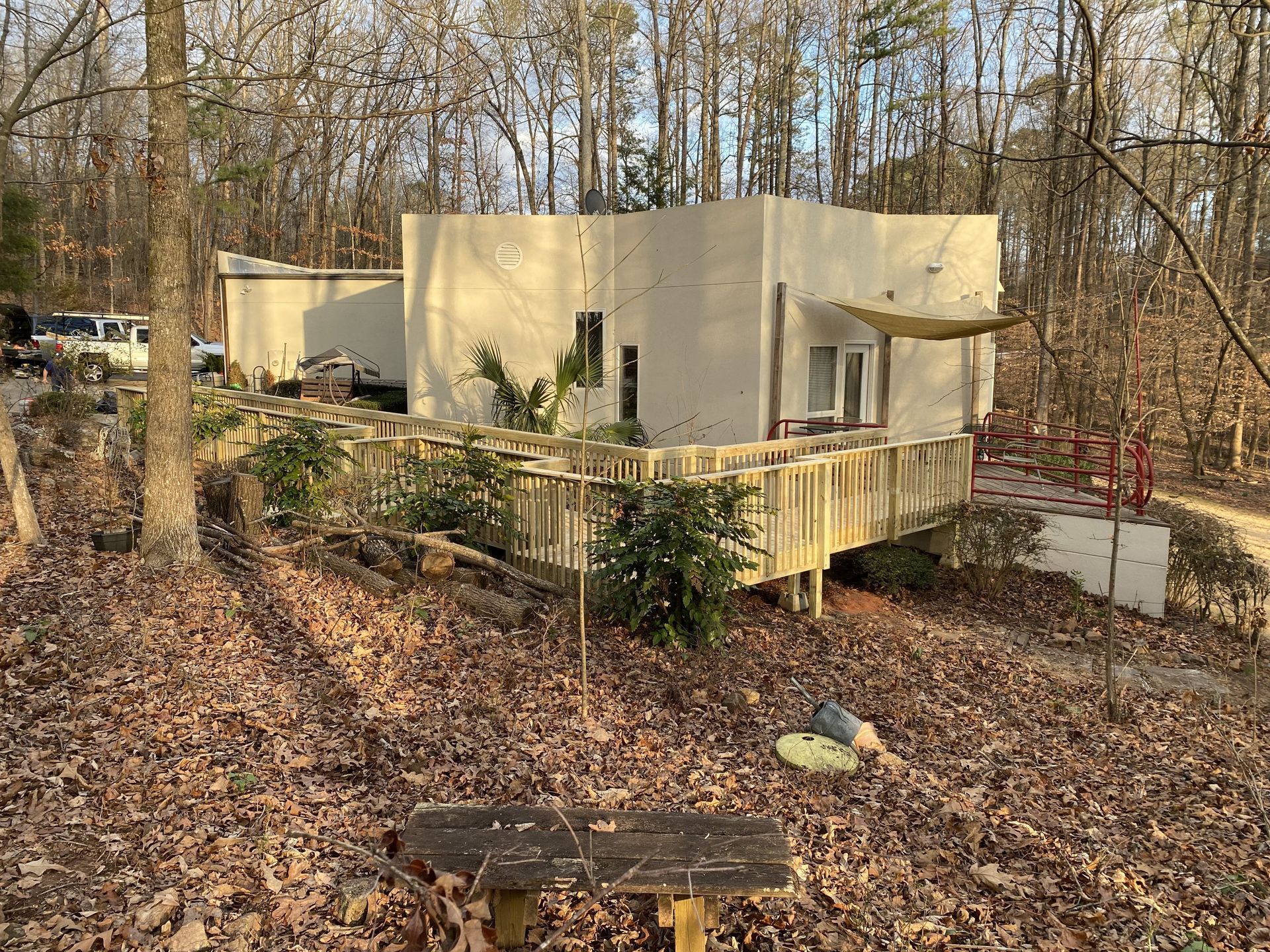 A small, light-colored building with a wooden deck in a wooded area. Brown leaves cover the ground.