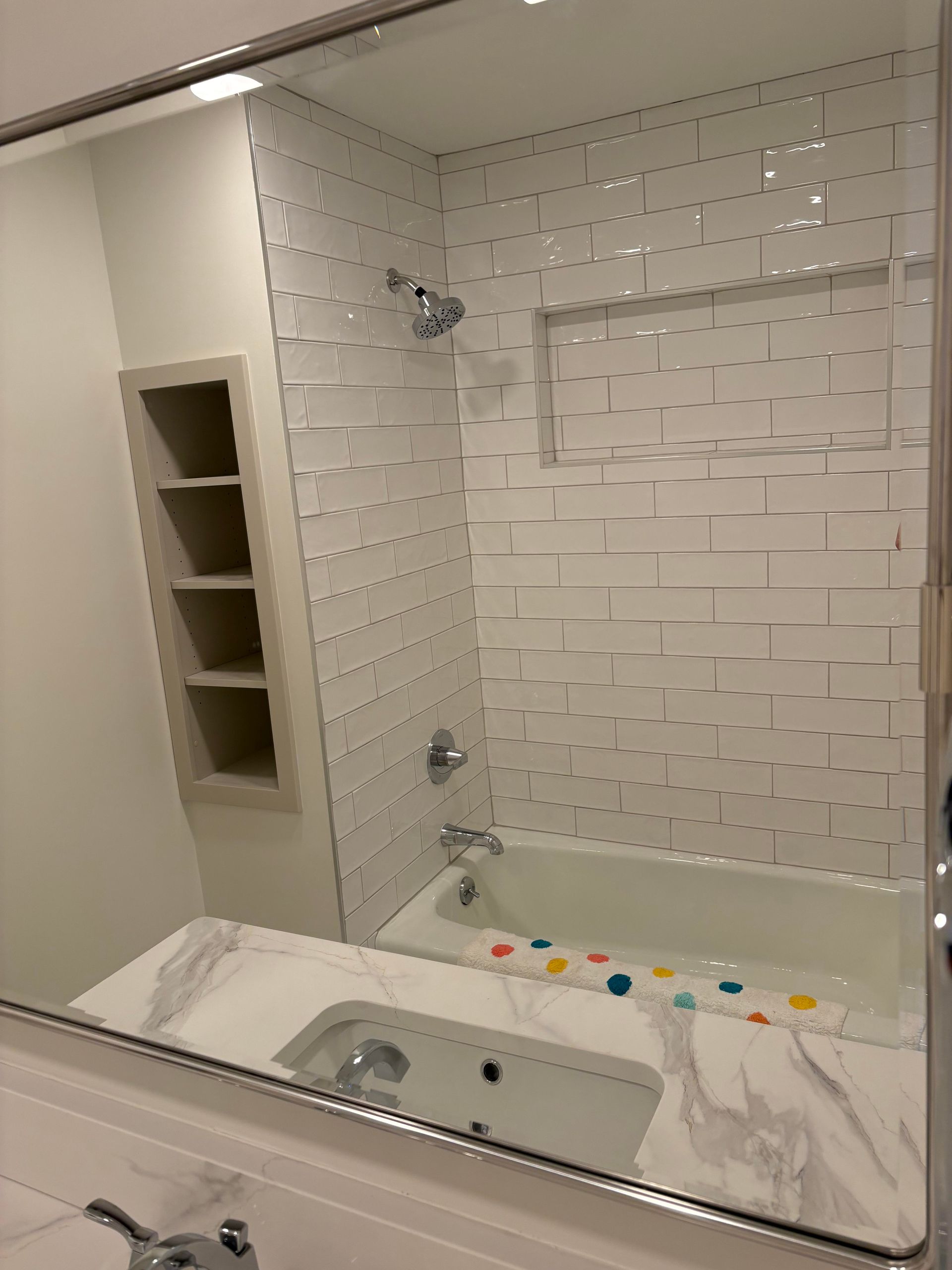 Bathroom with white tile shower, built-in shelving, white bathtub, and marble countertop.