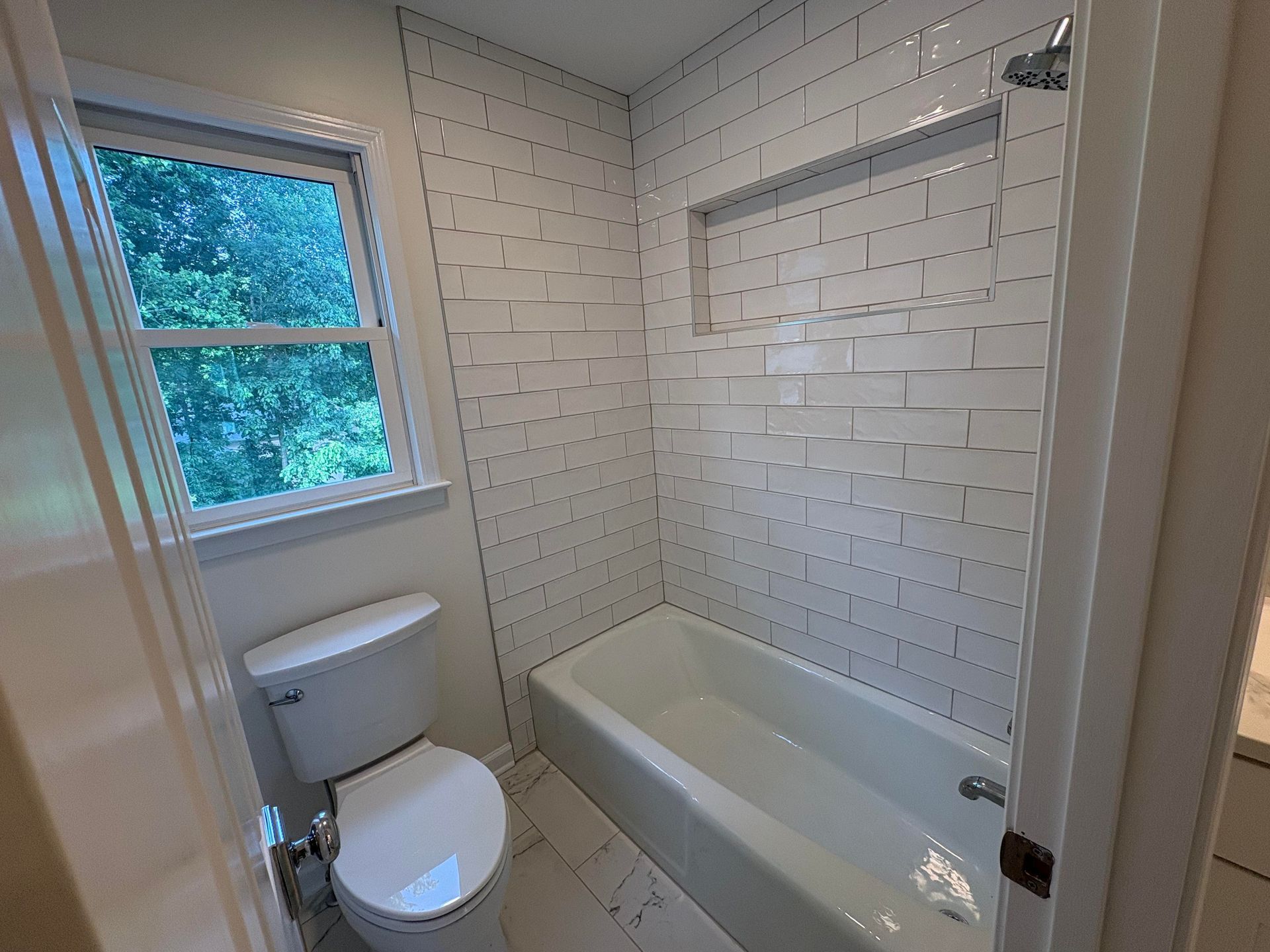 Bathroom with white subway tile, a bathtub, toilet, and window.