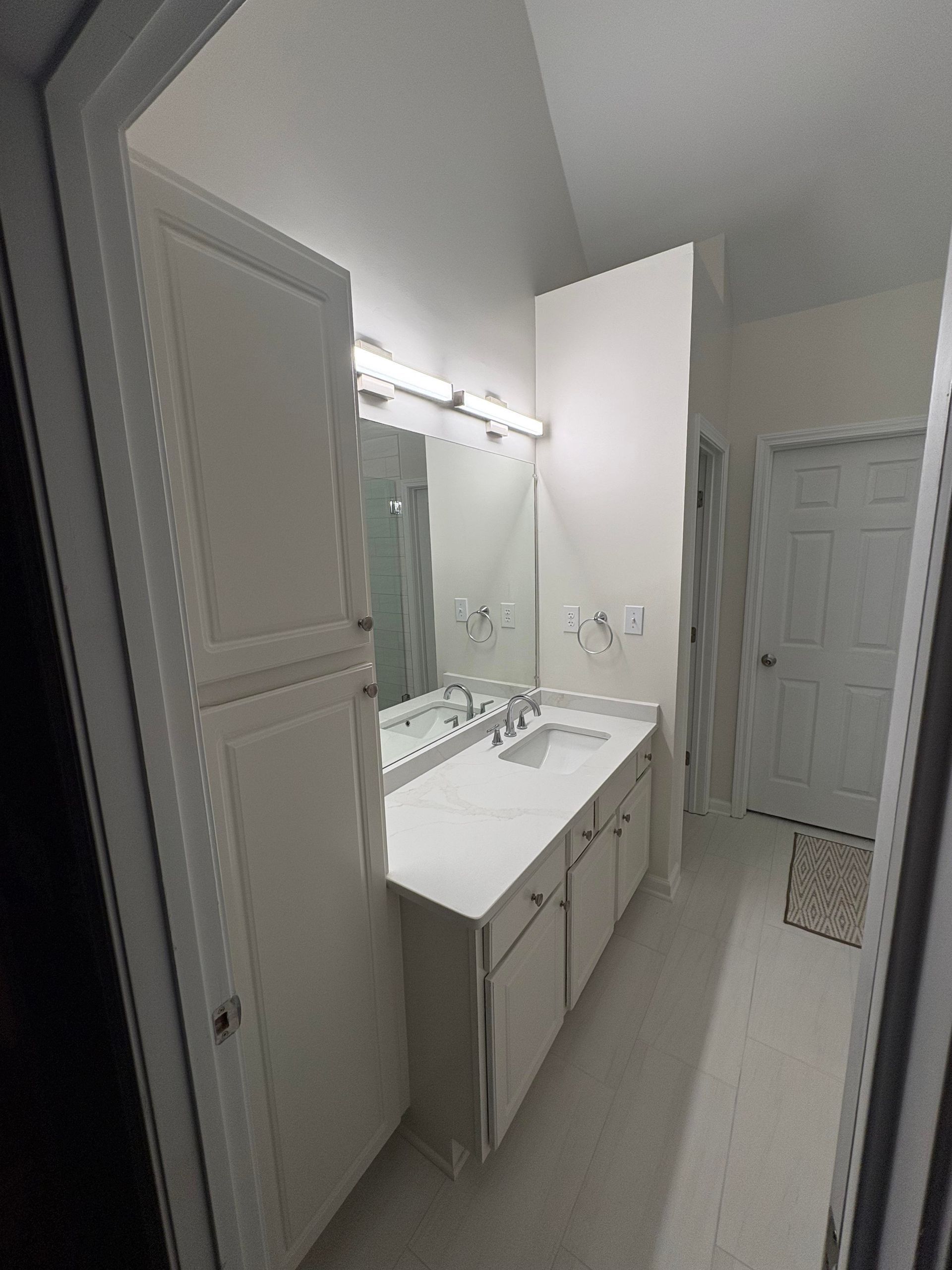 White bathroom with a vanity, mirror, and storage cabinet. A doorway leads to a closed door.