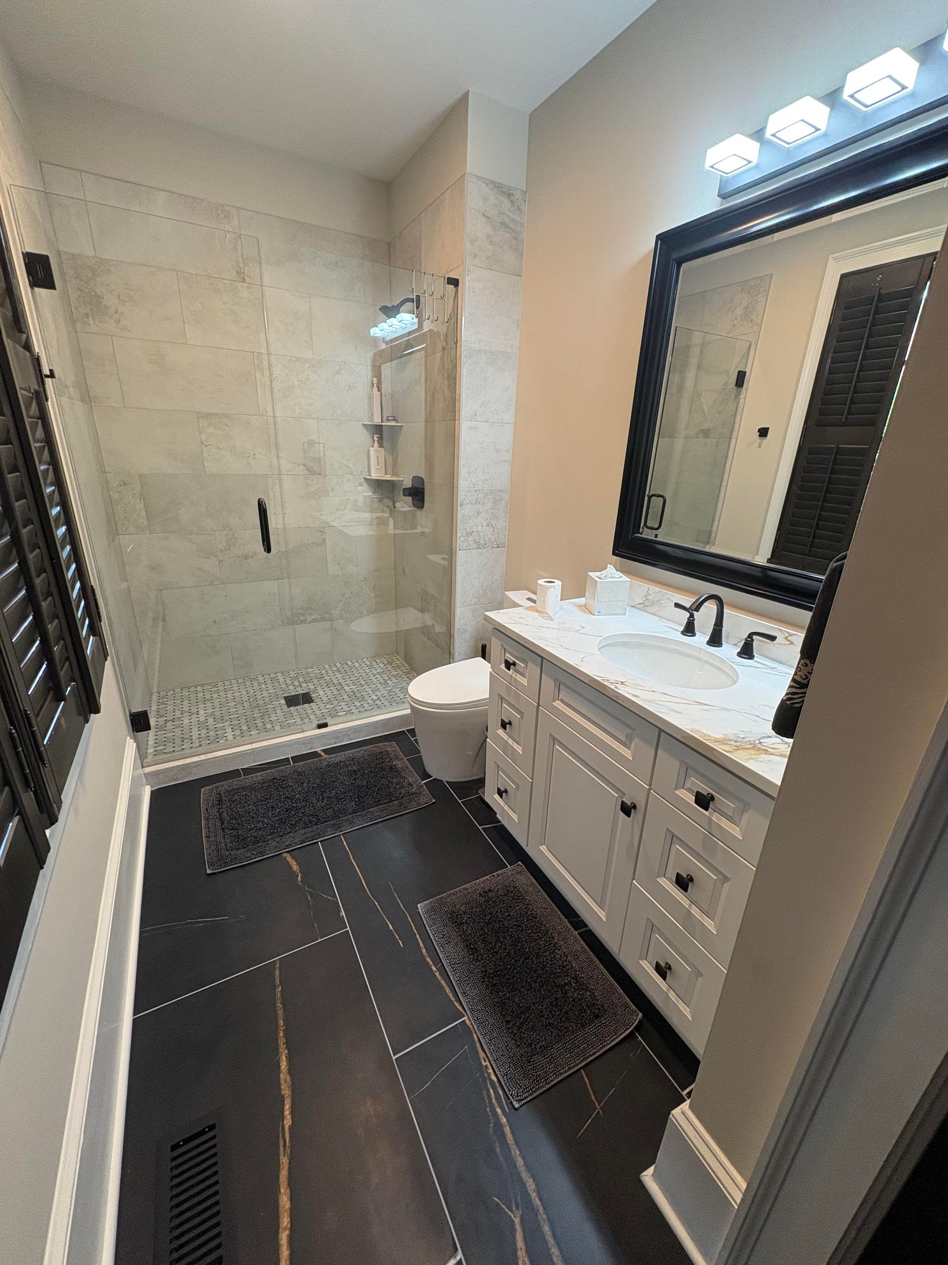 Bathroom with a white vanity, black framed mirror, and a glass shower. Black floor tiles.