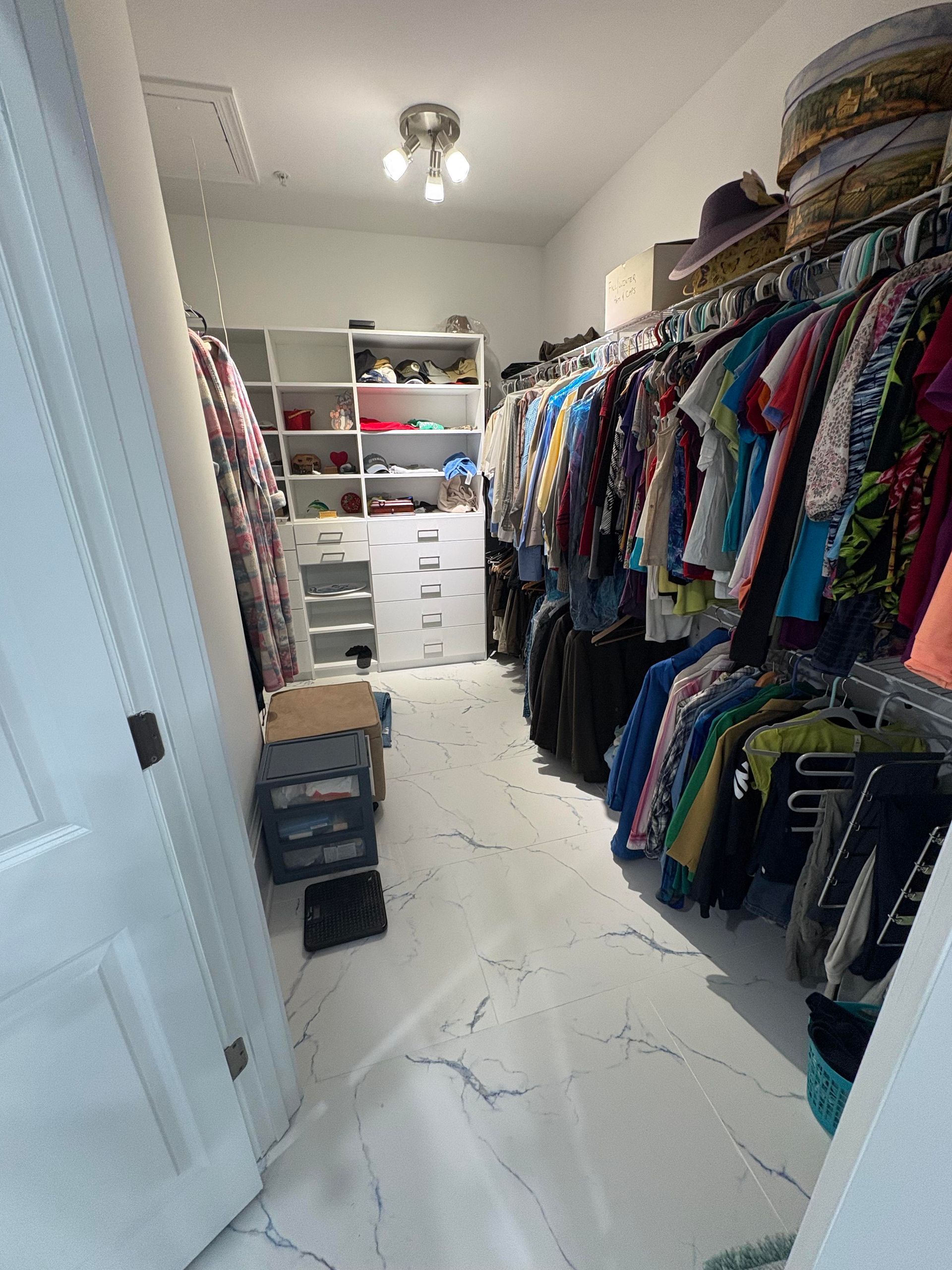 Walk-in closet with white shelving, a full rack of clothing, and white-tiled floor.