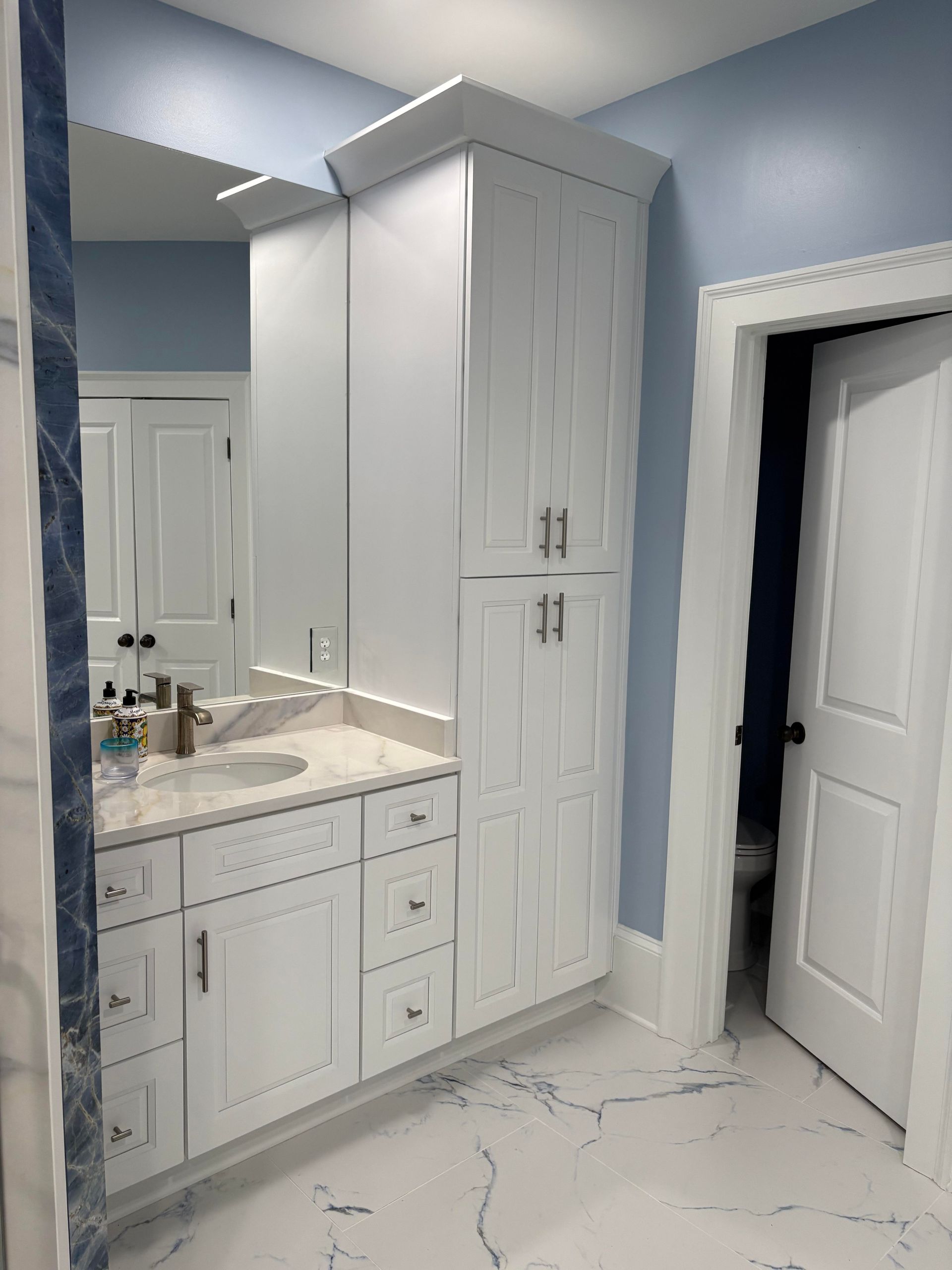 White bathroom vanity and tall cabinet against a light blue wall. White marble-look tile floor and open doorway.