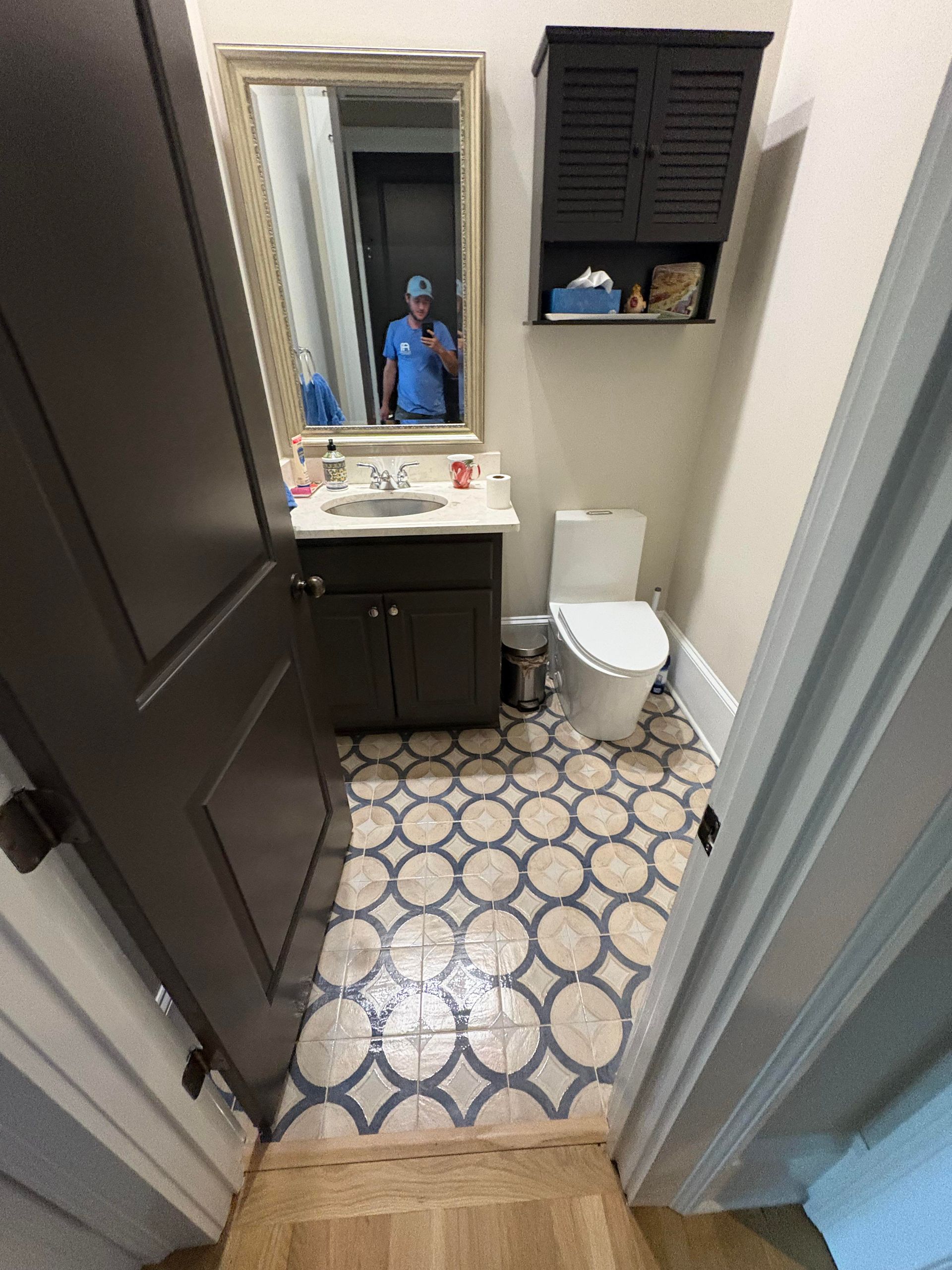 Small bathroom with patterned floor, dark cabinets, and a white toilet. A person is reflected in the mirror.