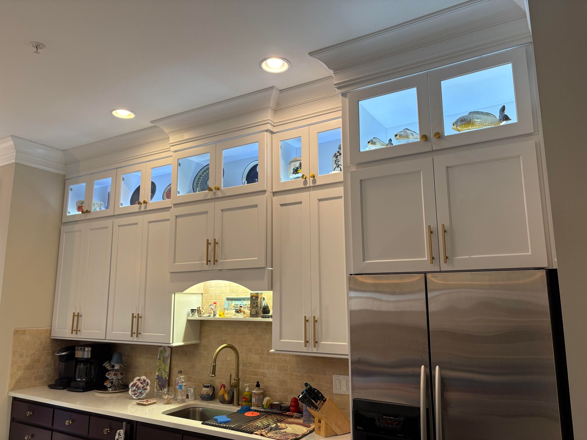White kitchen cabinets with glass-front display uppers, lit from within, over a countertop with sink and refrigerator.