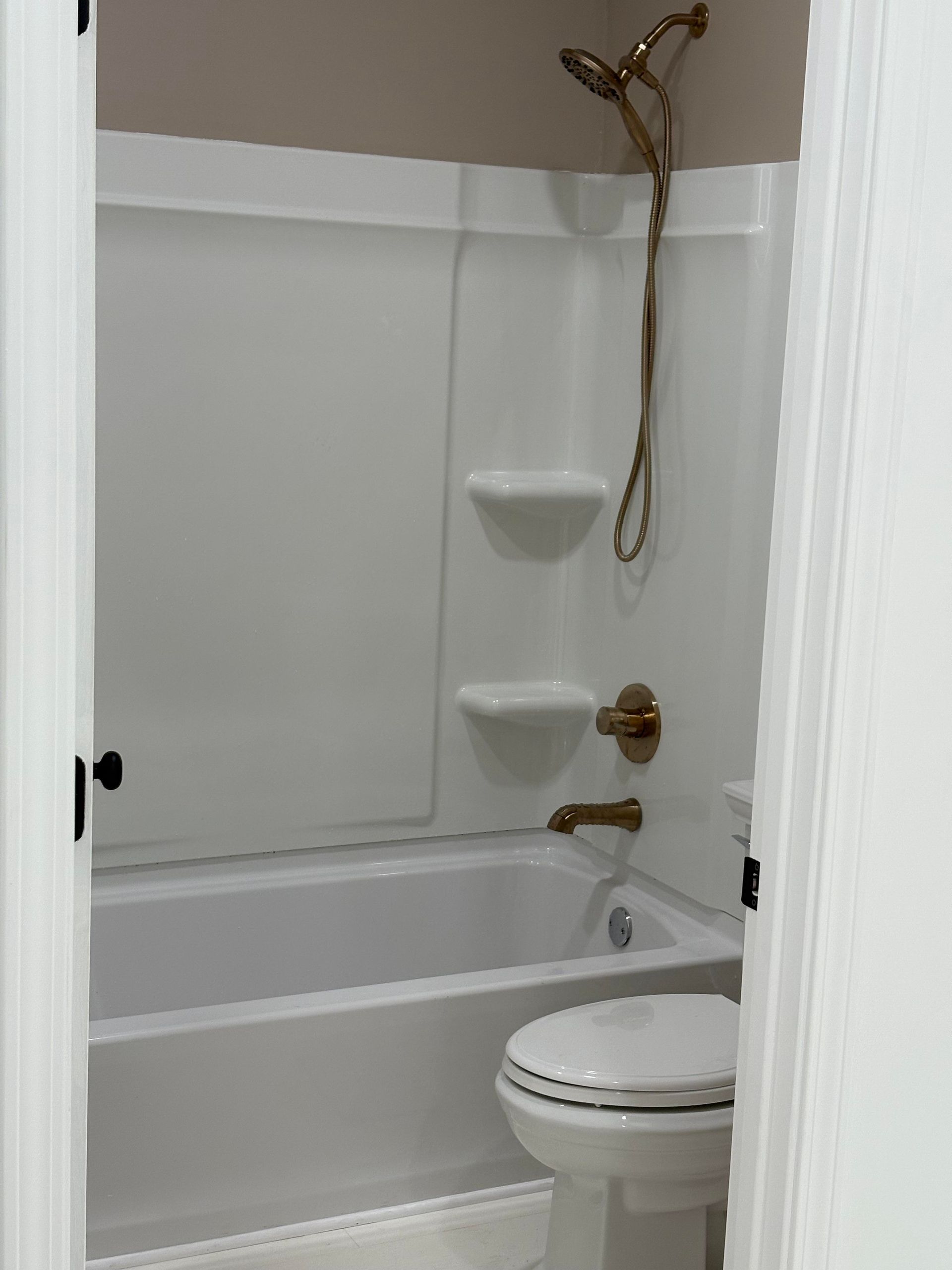 Bathroom interior: white tub and shower enclosure with bronze fixtures, toilet, and tan wall.