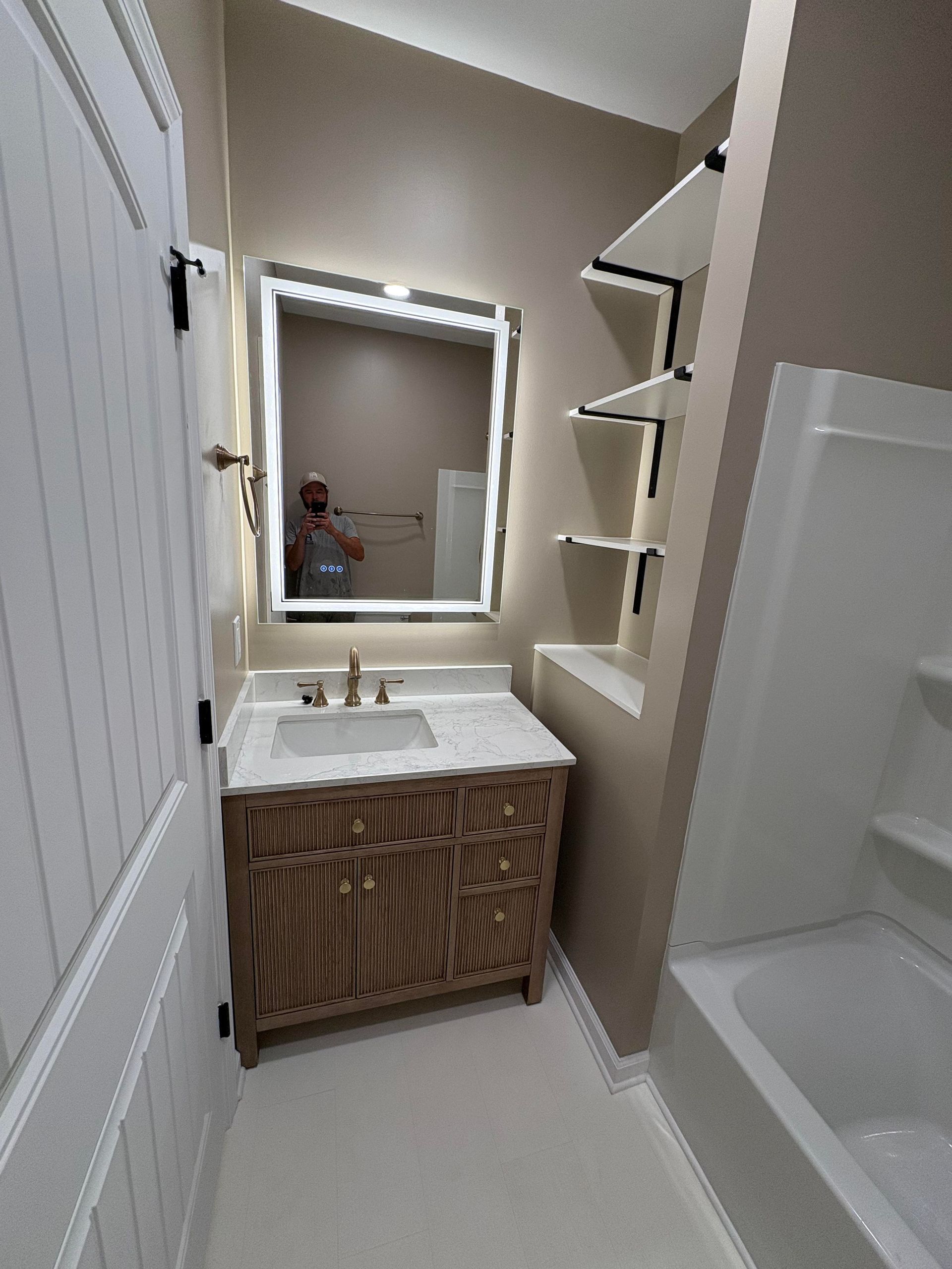 Bathroom with wood vanity, lit mirror, shelves, and tub. Light beige walls and white trim.