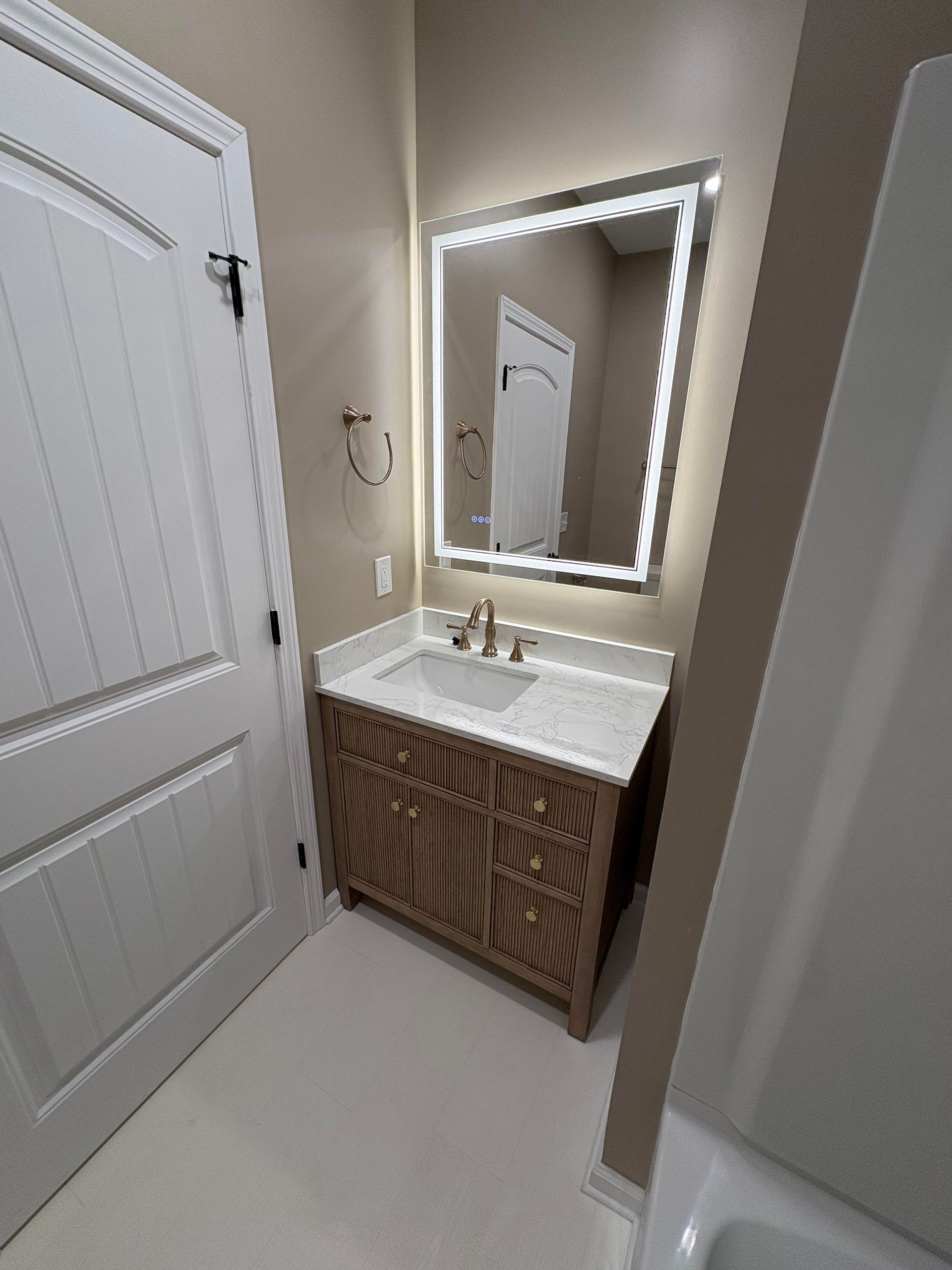 Bathroom with a vanity, lit mirror, white door, and a glimpse of a bathtub.