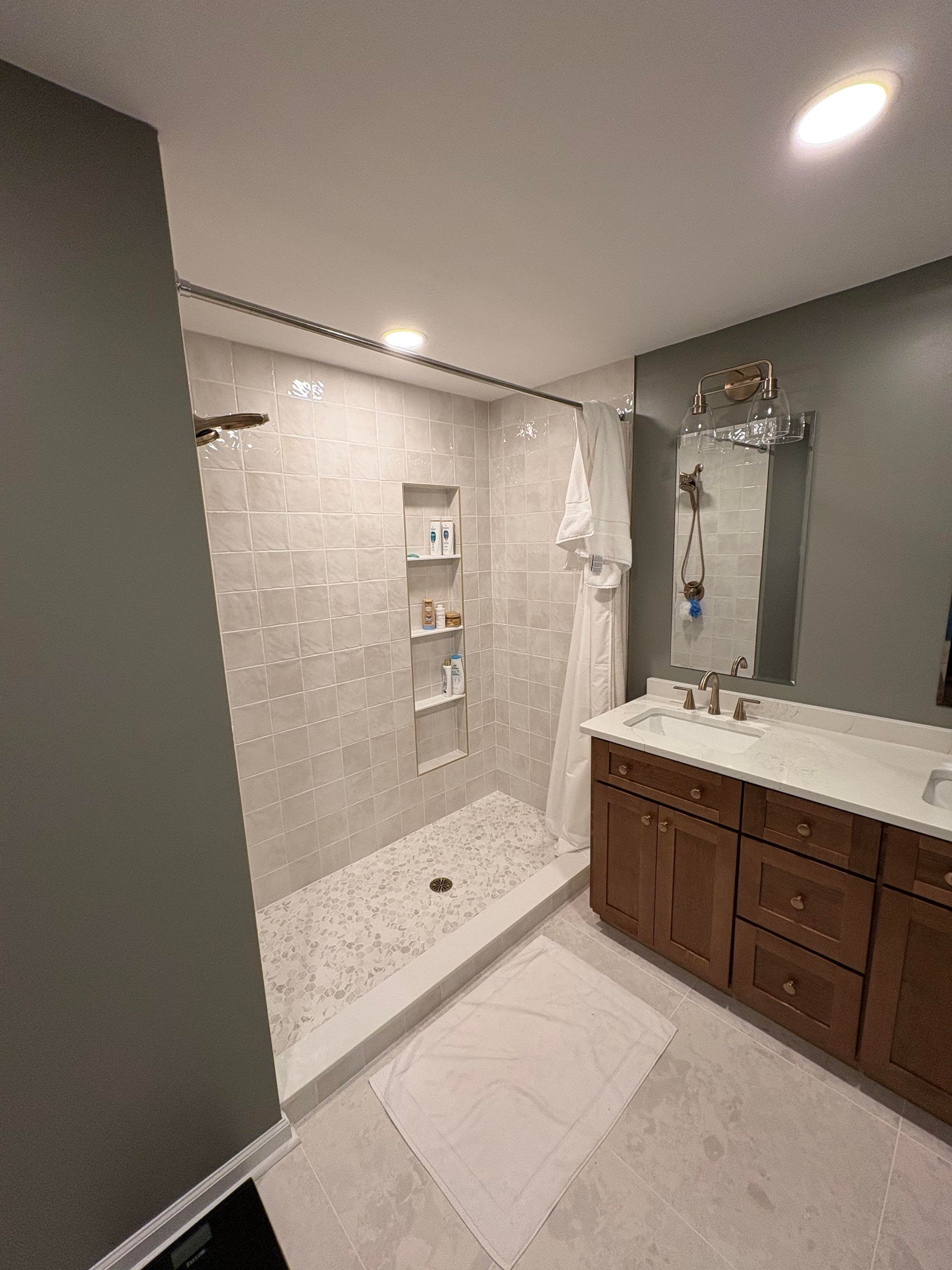 A modern bathroom with a walk-in shower. Gray walls, light gray tile, brown vanity.