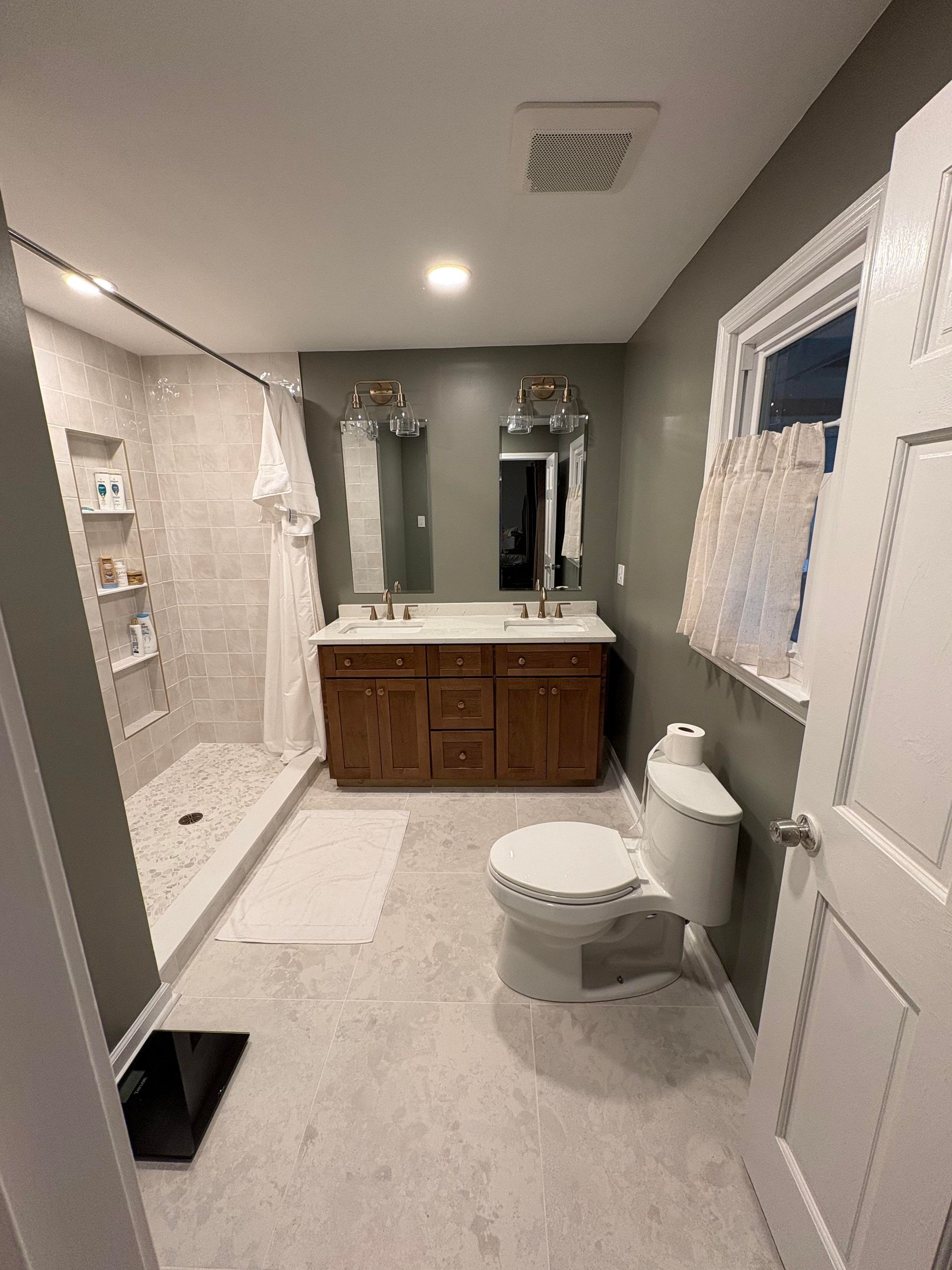 Bathroom with a walk-in shower, double vanity, and toilet; gray and brown tones, bright lighting.