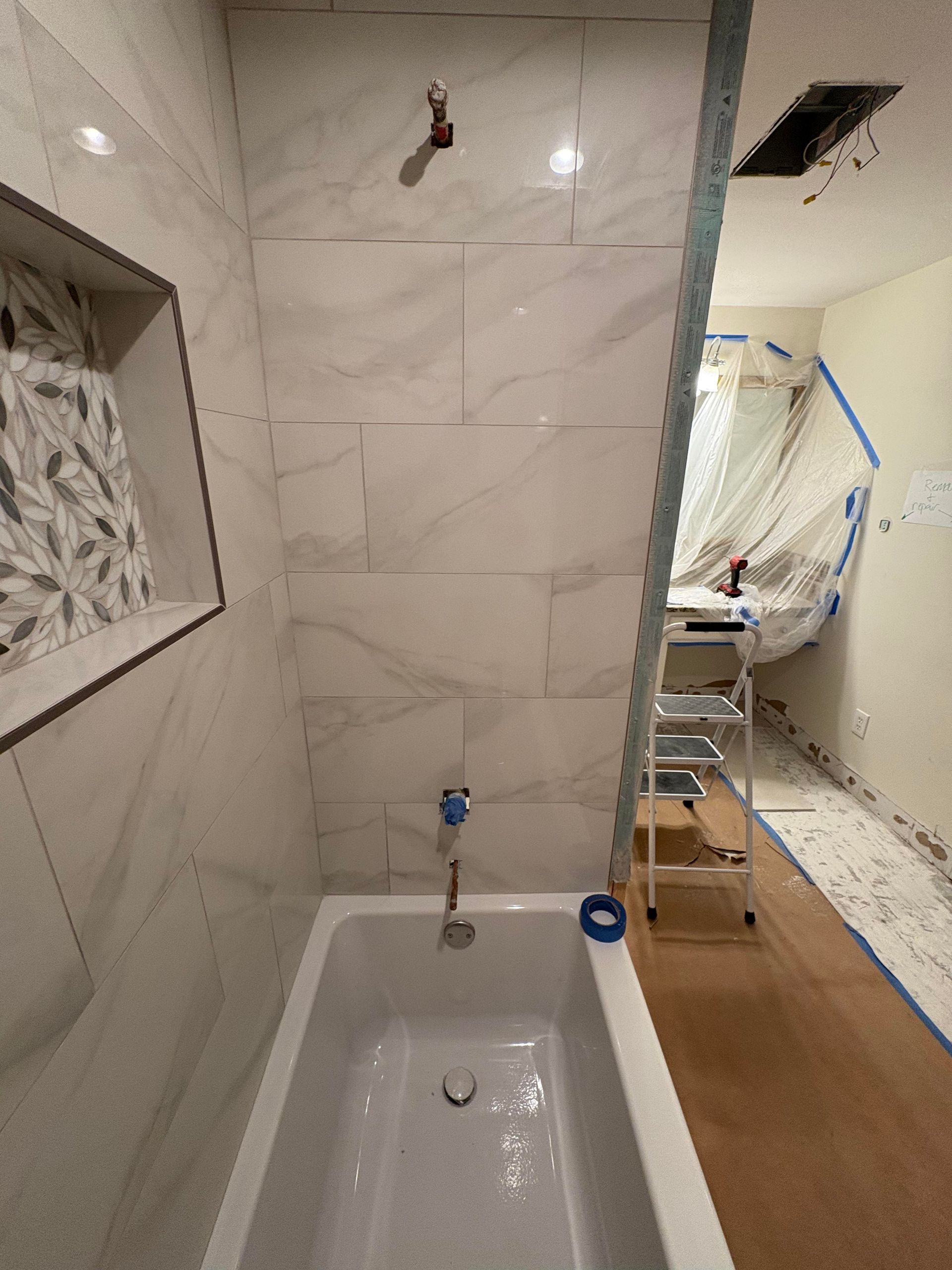 Bathroom under renovation, with tiled shower walls, bathtub, and exposed studs next to ladder and tarp.